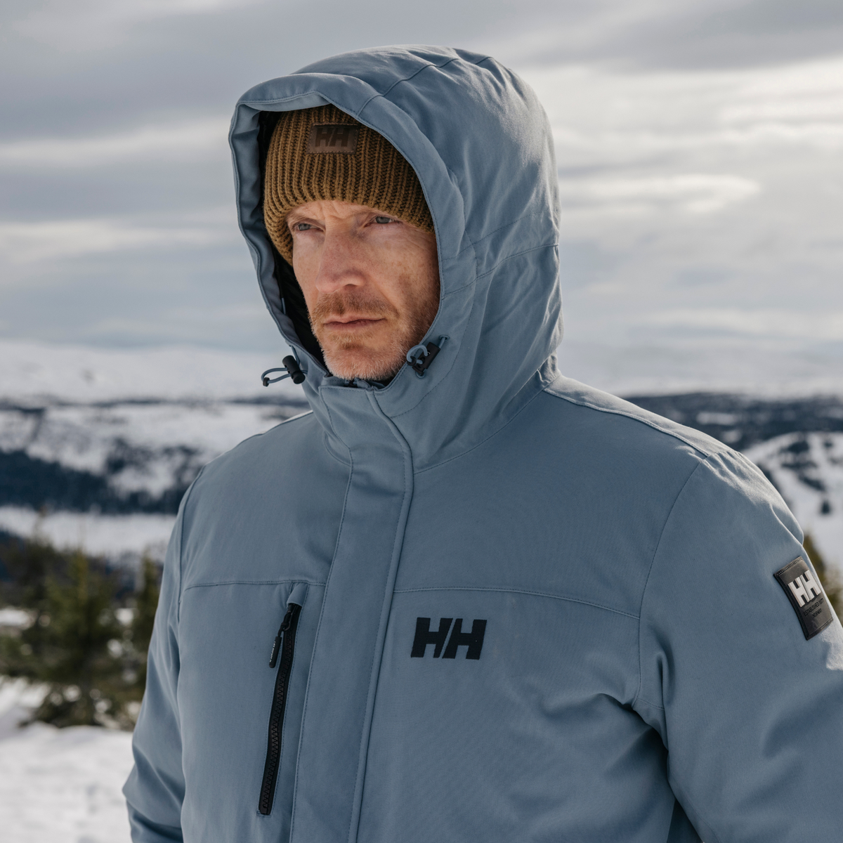 Man wearing a blue Helly Hansen jacket with a Helly Hansen Unisex HH Rib Beanie Hat Sepia 718 54020 in a snowy landscape