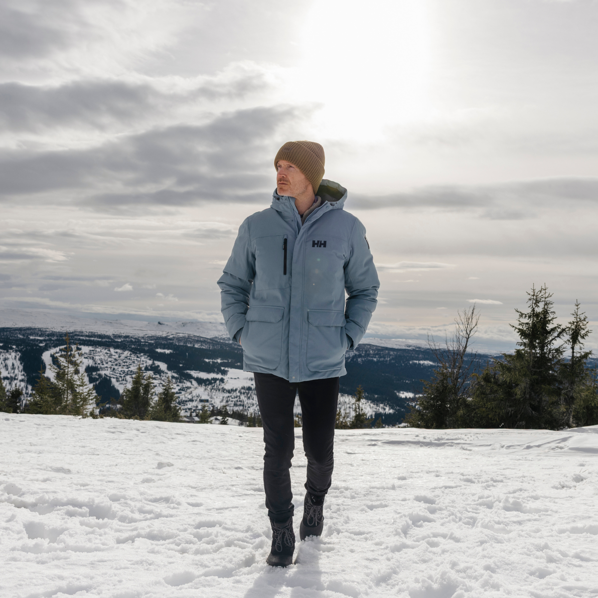 Person wearing a blue jacket with a Helly Hansen Unisex HH Rib Beanie Hat Sepia 718 54020 standing on a snowy landscape with mountains in the background