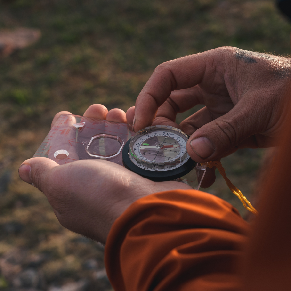 A close-up of a person's hands holding a Highlander Deluxe Map Compass COM006, outdoors with a blurred natural background.