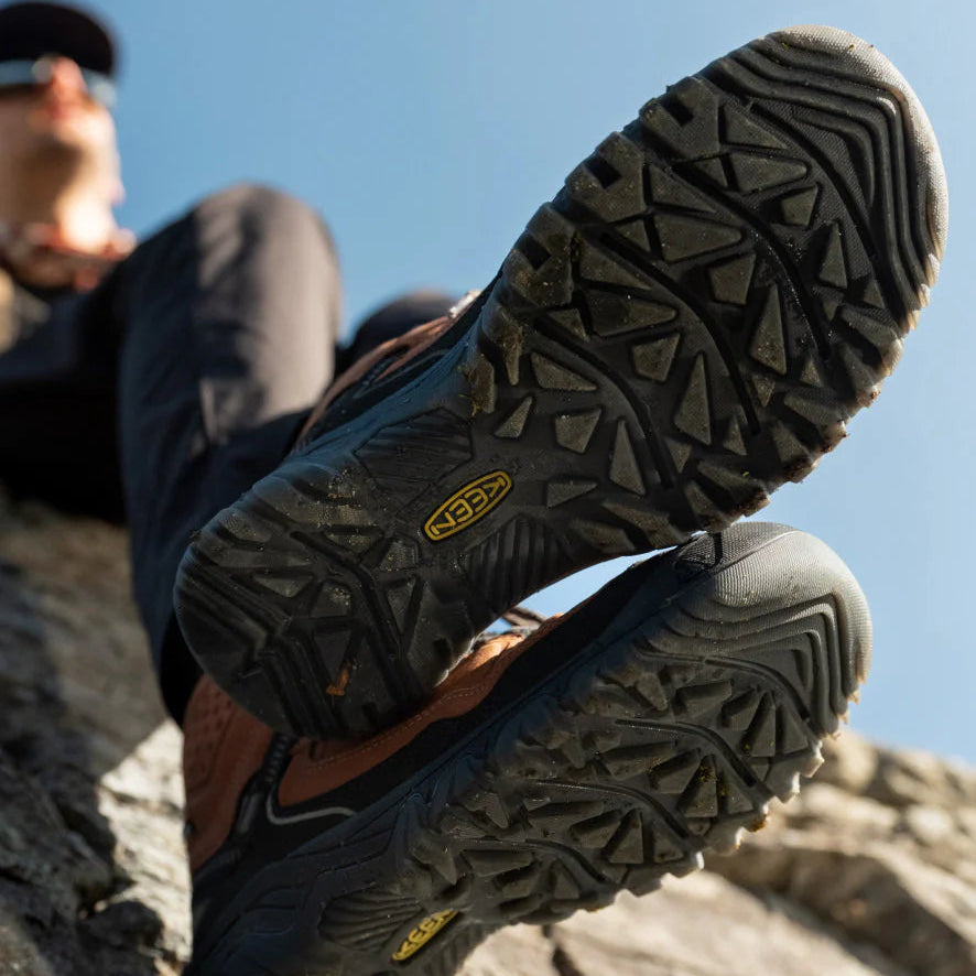 Person wearing Keen Men's Targhee IV Waterproof Mid Trail Boots Wide Fit Bison Black 1029002, showing the soles of the shoes, sitting on a log with a rocky outcrop with a blue sky background