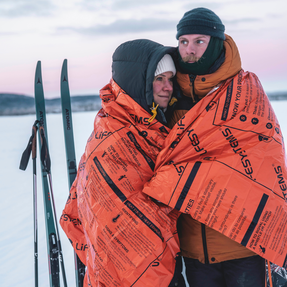 Two people in a snowy setting, with skis in the background, with a Lifesystem Heatshield Survival Blanket Double 42170 wrapped around both of them