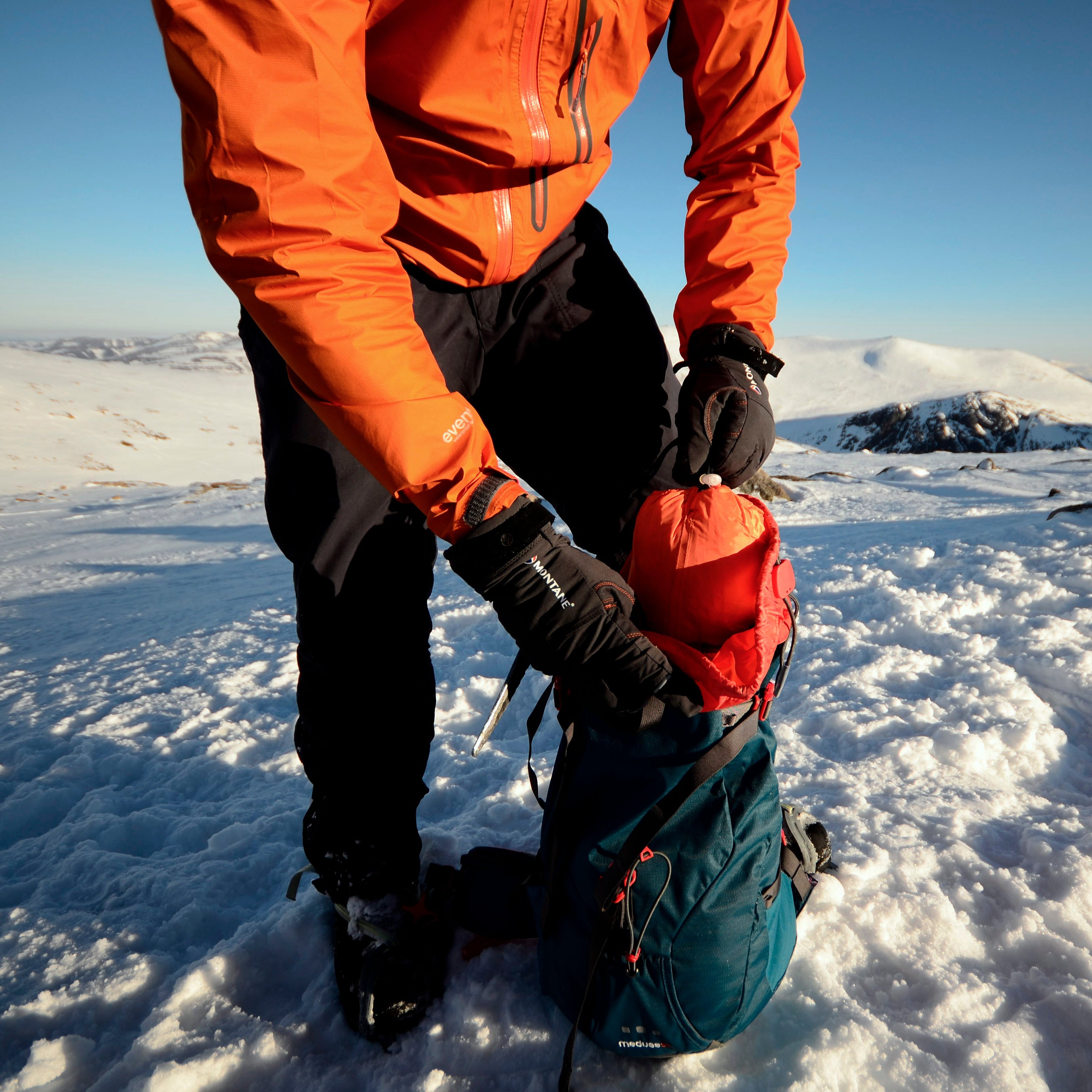Person in orange jacket and black trousers taking a Lifesystems Survival Shelter 2 People Orange 42311 out of their pack on a snowy mountain