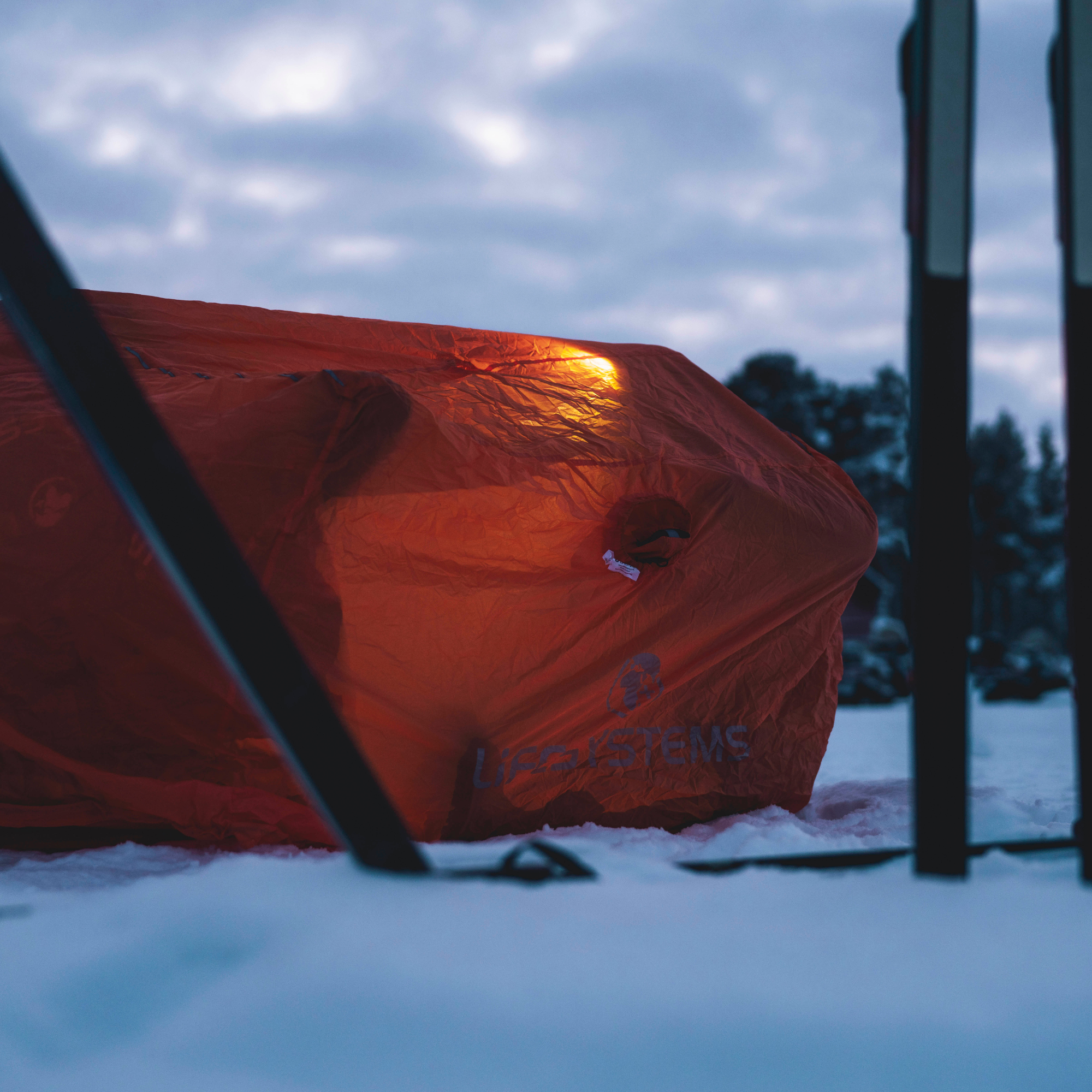 A Lifesystems Survival Shelter 4 People Orange 42321 in snow outdoors at dusk, with a warm light glowing from inside and blurred dark poles in the foreground.