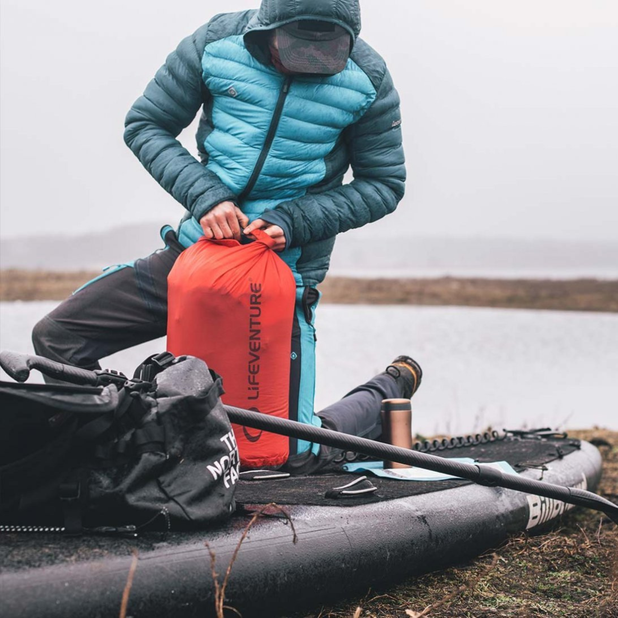 Person on a stand up paddle board holding a Lifeventure Ultralight Dry Bag with a lake in the background
