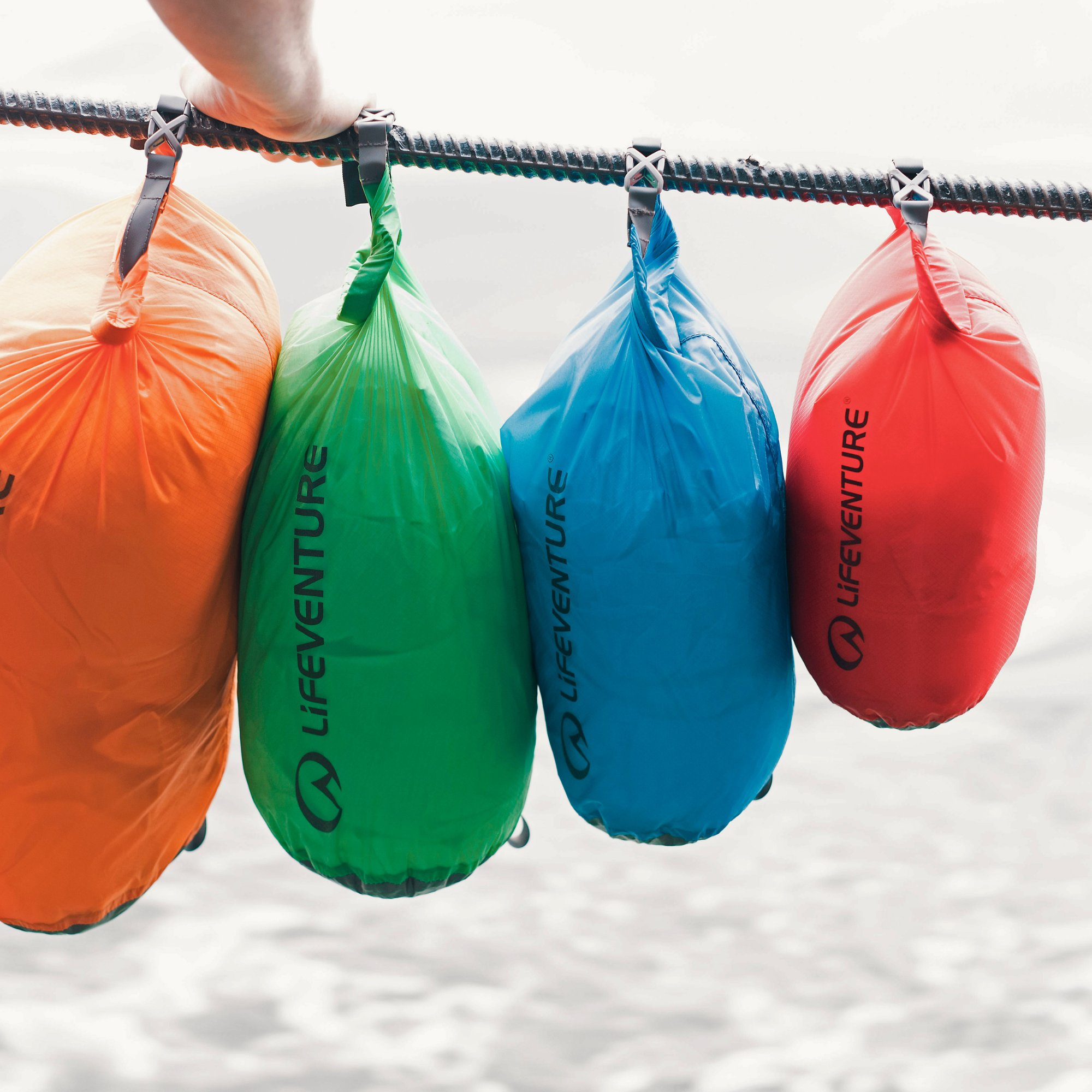 Four Lifeventure waterproof Ultralight Dry Bags in orange, green, blue, and red hang from a rope outdoors.