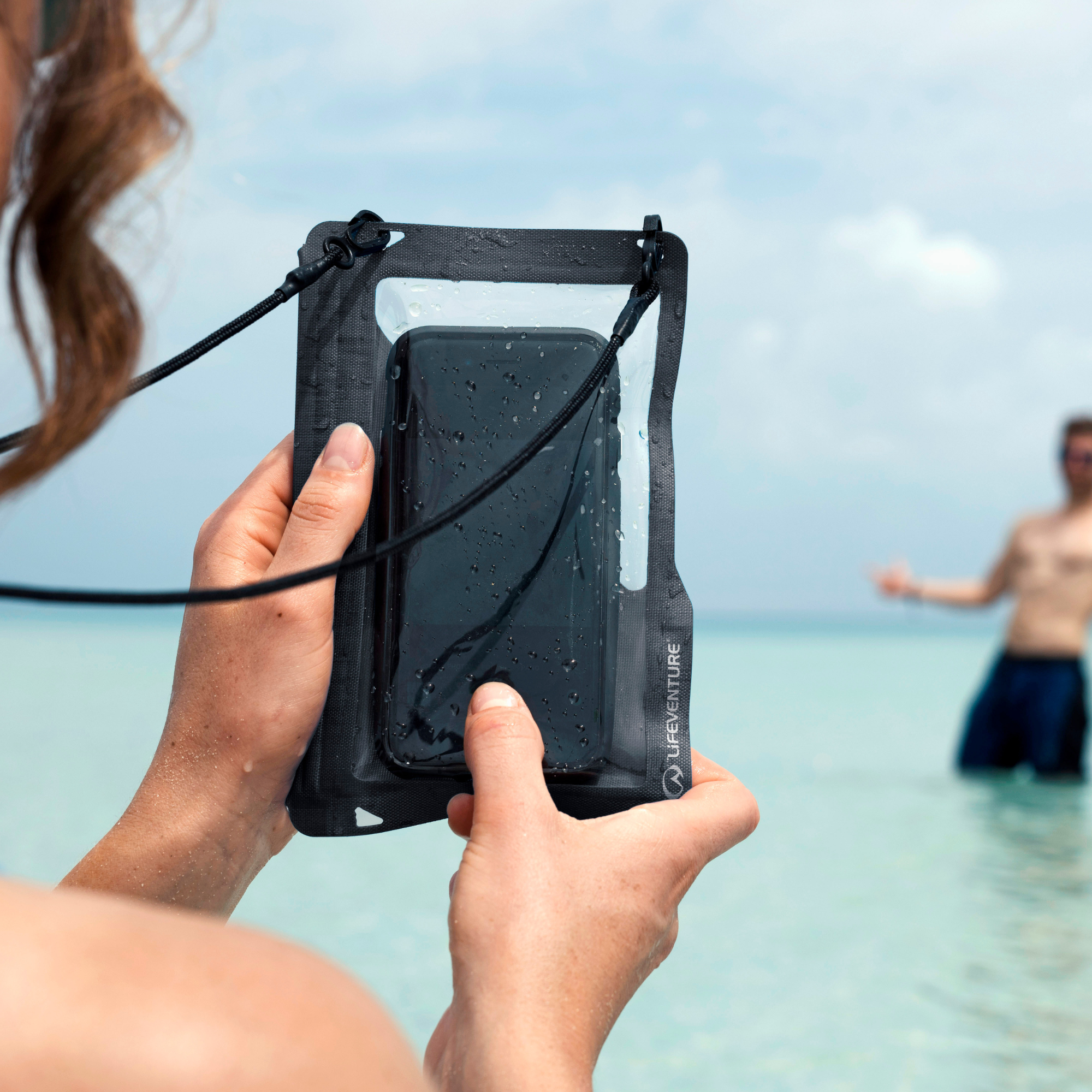 A person holding up a Lifeventure Waterproof Phone Case Grey 59551 with a clear front window at a beach, taking a photo of a man standing in the ocean water blurred in the background.