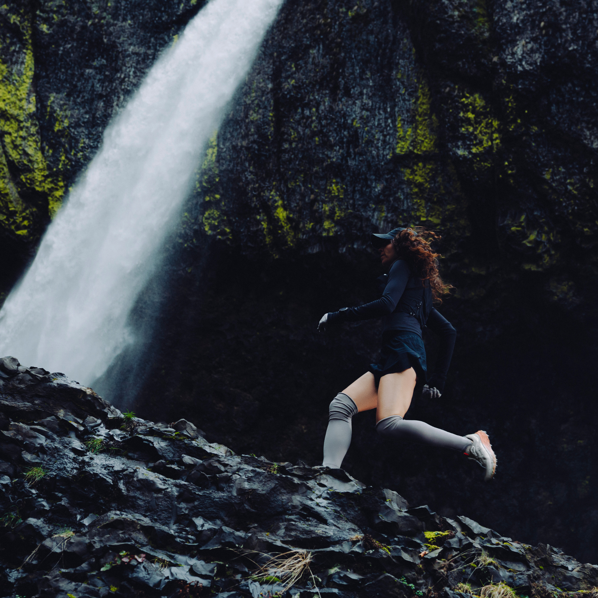 A woman in a black athletic outfit, grey knee-high socks and Merrell Women's Agility Peak 6 Vegan Gore-Tex Trail Running Shoes Diorite J00005818 runs along a rocky path with a large waterfall cascading down a mossy cliff behind her.