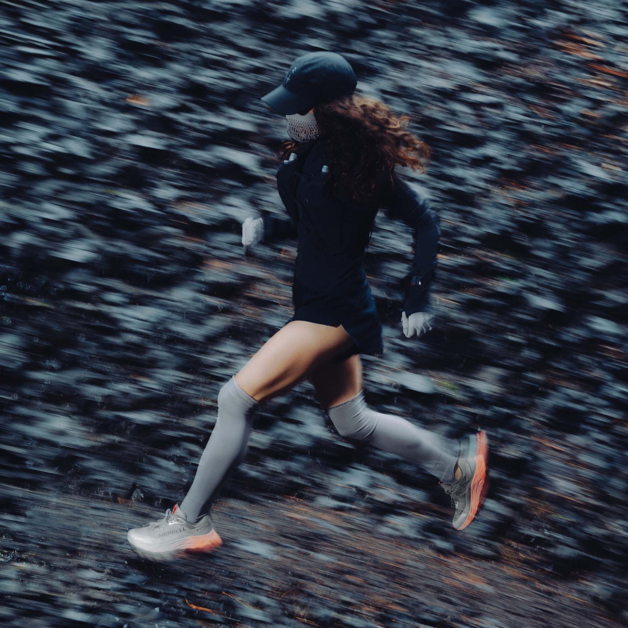 A woman running outdoors wearing a black long-sleeve top, black shorts, grey knee-high socks, and Merrell Women's Agility Peak 6 Vegan Gore-Tex Trail Running Shoes Diorite J00005818, captured with a motion-blur effect against a dark forest background.