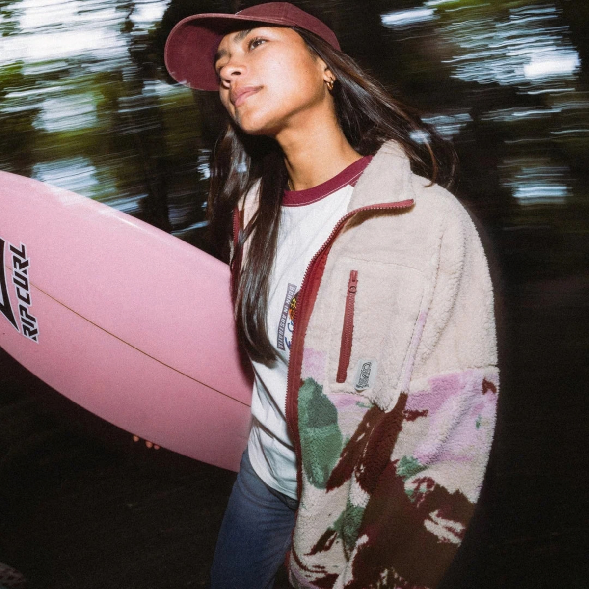 A woman wearing a Rip Curl Women's Search Series Bonded Polar Full Zip Fleece Jacket Dark Plum 2924 0DDWFL, a white t-shirt, and a maroon baseball cap, holding a pink surfboard. The background is a dark, blurred outdoor scene with motion blur.