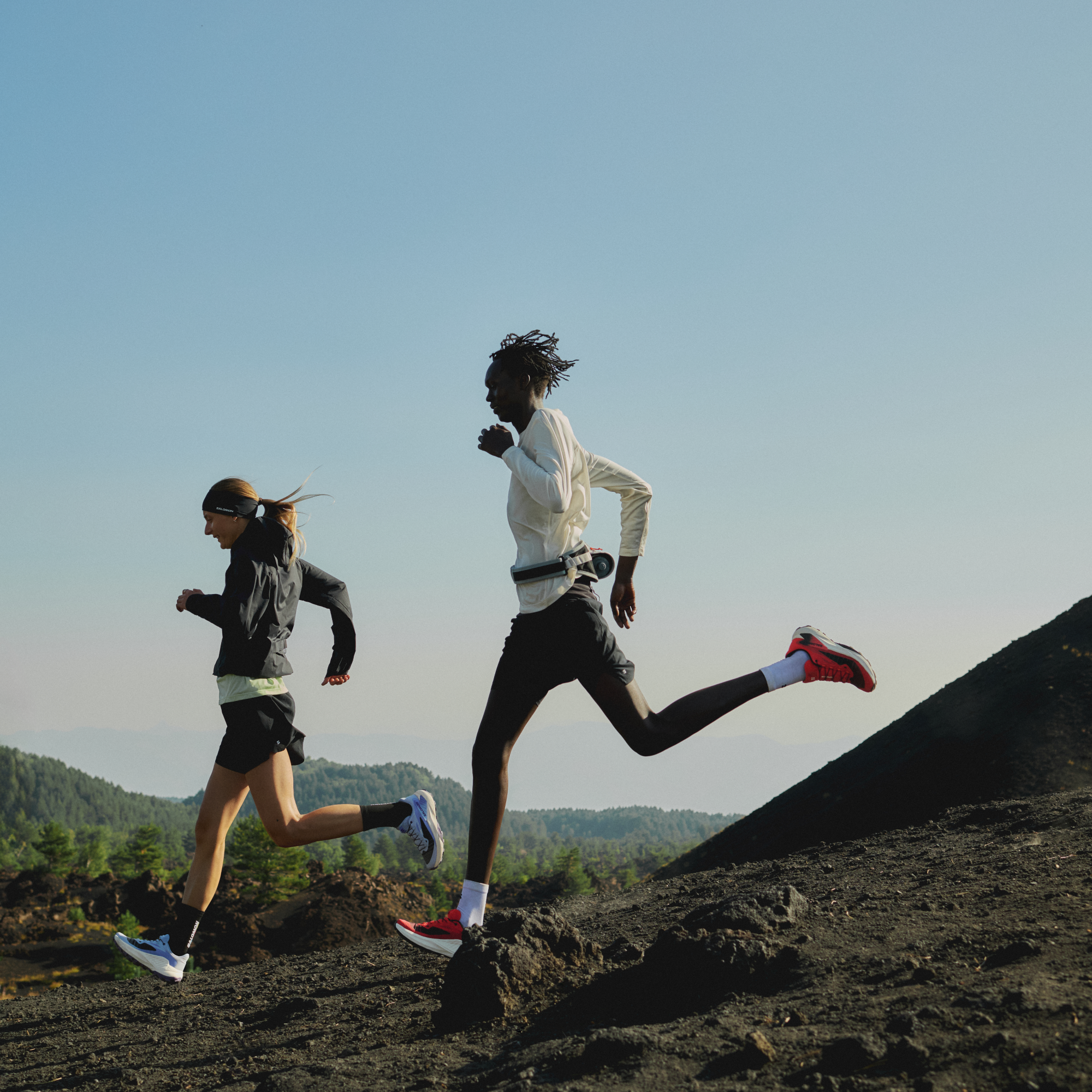 2 people running on a rocky landscape. Man wearing Salomon Men's Genesis Trail Running Shoes Fiery Red Lava Falls Black L49226500