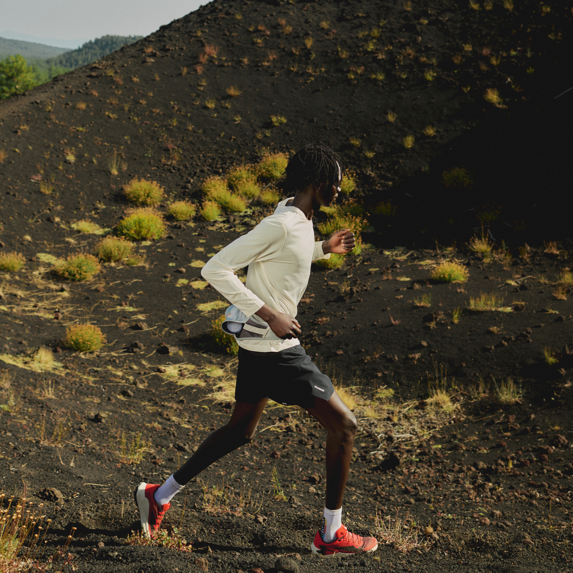 Person running on a rocky hillside with a clear sky wearing Salomon Men's Genesis Trail Running Shoes Fiery Red Lava Falls Black L49226500