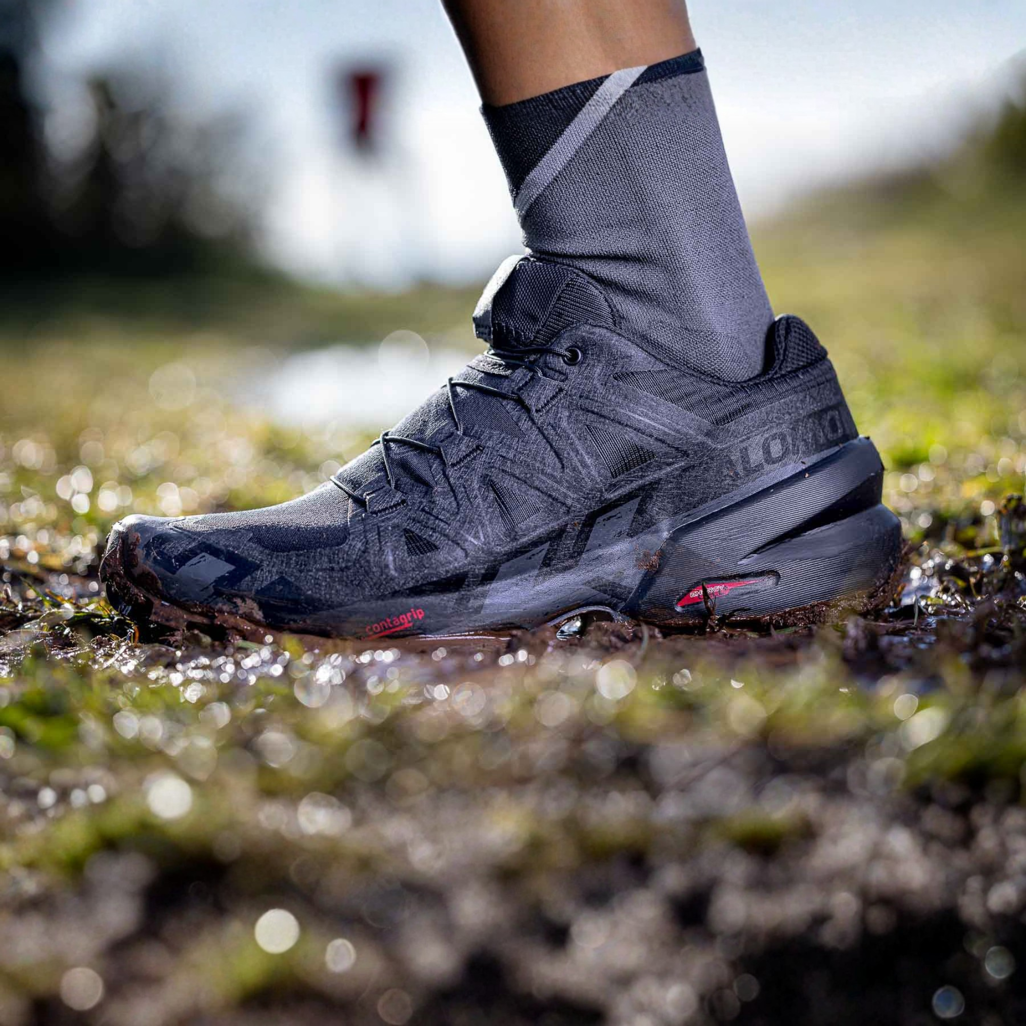 A close-up shot of a person's foot wearing a Salomon Men's Speedcross 6 Trail Running Shoes Black Phantom L41737900 and a grey sock, standing in shallow water on a muddy, grassy outdoor ground.