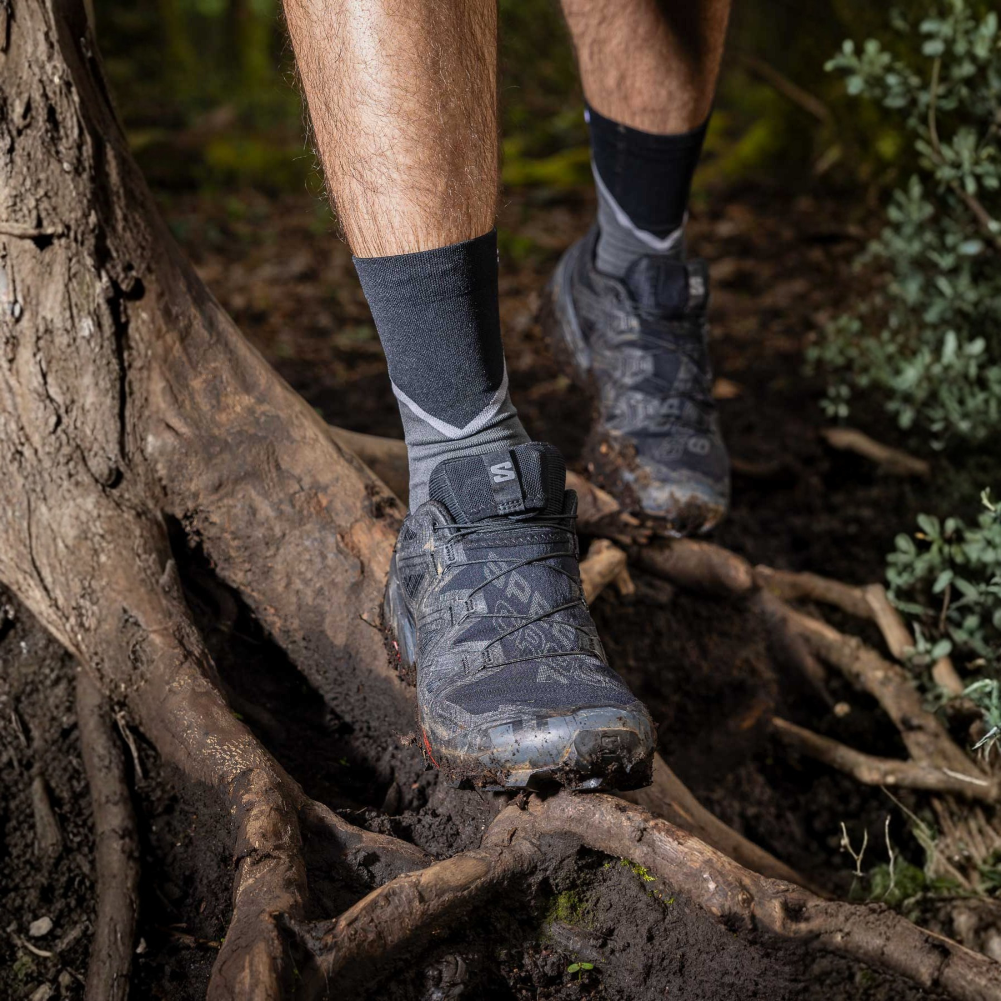 A close-up outdoor shot of a person trail running through mud on a forest trail, wearing Salomon Men's Speedcross 6 Trail Running Shoes Black Phantom L41737900 and dark grey crew socks.