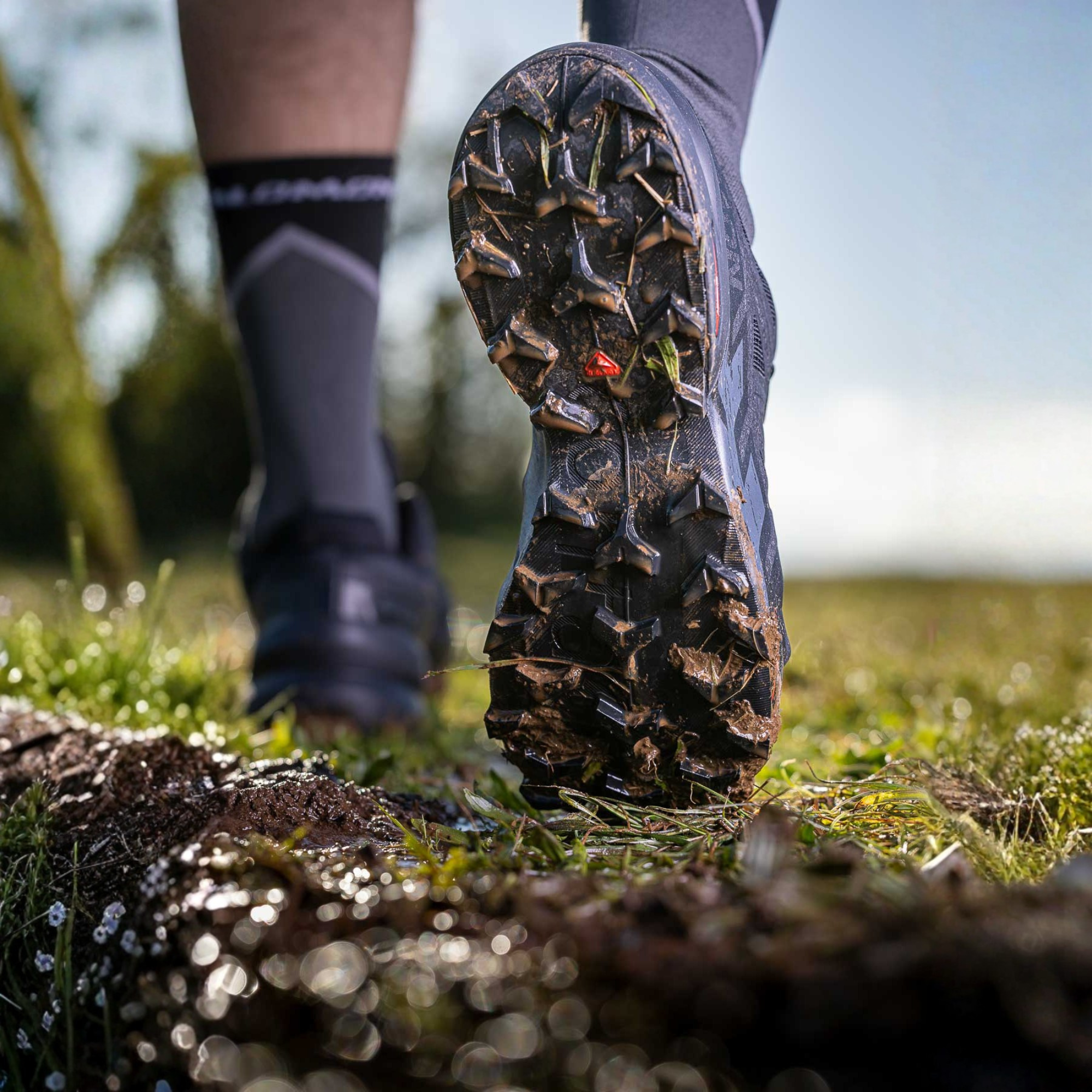 A close-up action shot of a trail runner's foot pushing off from a muddy trail, wearing Salomon Men's Speedcross 6 Trail Running Shoes Black Phantom L41737900 showing the rugged black tread with a tiny red logo. The runner is wearing dark grey socks and another shoe is visible in the background on a grassy outdoor trail.