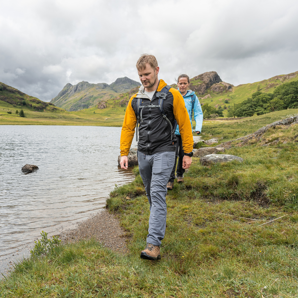 A man hiking along the grassy shore of a lake, wearing a black and yellow outdoor jacket with Scarpa Men's Cyrus 2 Gore-Tex Trail Shoes Brown Black 63326-200, with a woman in a blue jacket following behind in a mountain landscape under a cloudy sky.