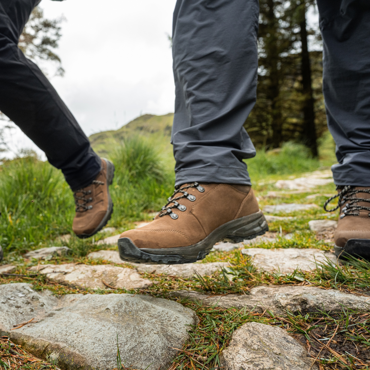 A close-up outdoor shot of a person hiking on a stone path, wearing Scarpa Men's Cyrus 2 Gore-Tex Trail Shoes Brown Black 63326-200, grey trousers, and another person's legs and boot are visible in the background.