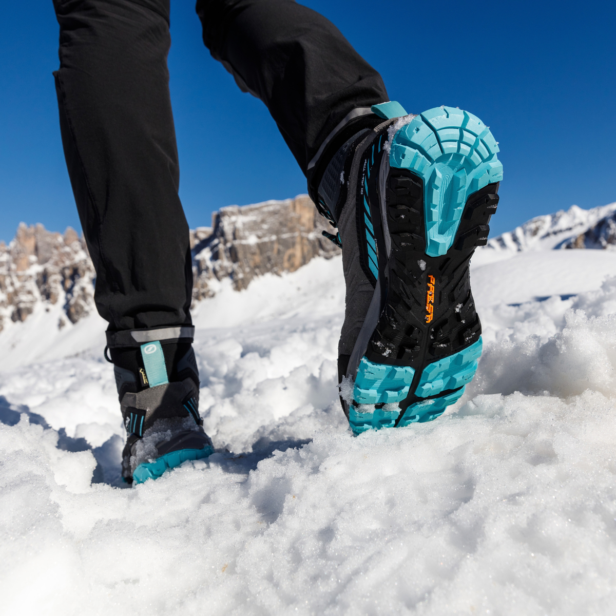 A close-up of a person's legs walking through deep snow, showcasing the Presa outsole of a Scarpa Women's Rush TRK Gore-Tex Hillwalking Boot Mid Grey Aqua 63143-202. The background features a bright blue sky and rocky mountains.