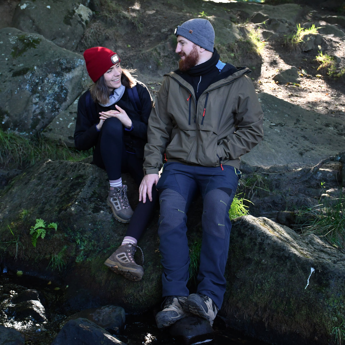 Two people sitting on rocks in a forested area beside a stream. Man wearing Six Peaks Unisex LED Lighted Beanie Hat Grey SIH0004