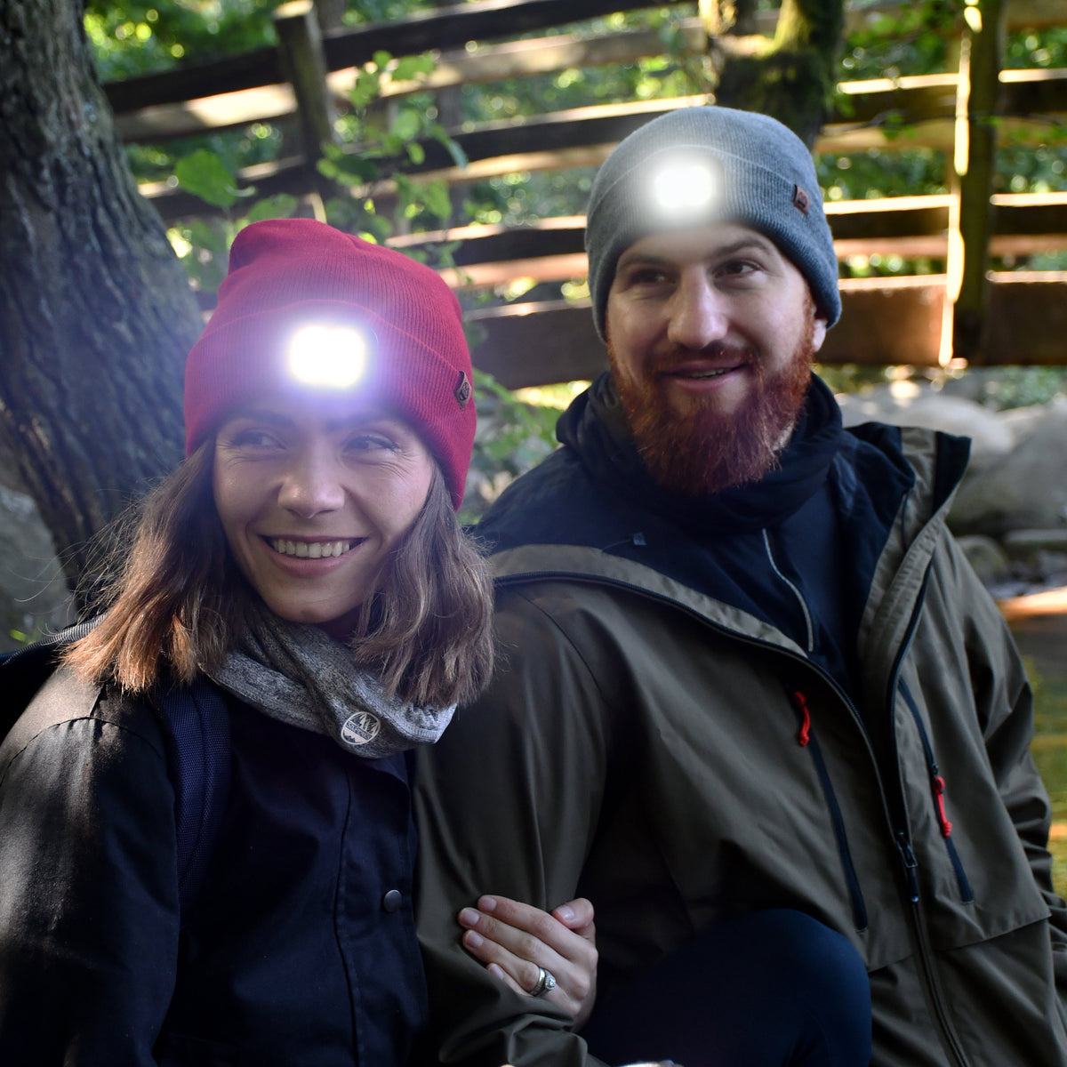 Two people sitting outdoors, surrounded by trees and a wooden fence. Man wearing Six Peaks Unisex LED Lighted Beanie Hat Grey SIH0004