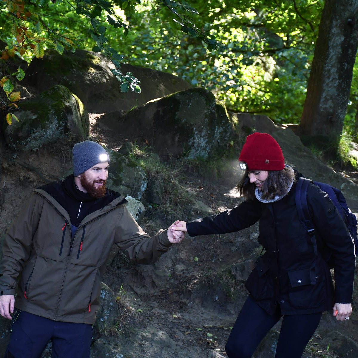 Two people in a forest setting, one wearing a red beanie and the other a Six Peaks Unisex LED Lighted Beanie Hat Grey SIH0004, with a backpack.