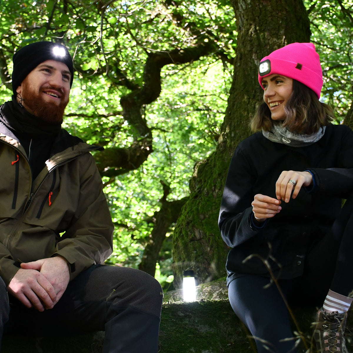 Two people sitting outdoors in a forested area, woman wearing Six Peaks Unisex LED Lighted Beanie Hat Pink SIH0004.