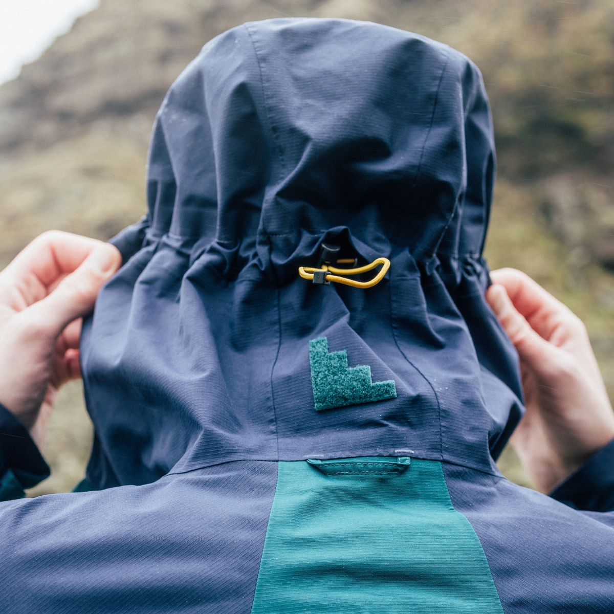 Close-up back view of a person wearing a Sprayway Men's Maxen 2L Gore-Tex Waterproof Jacket SP-01754 Dark Sea Blazer SP-008873, with both hands adjusting the hood. Set against a blurred natural mountain background.