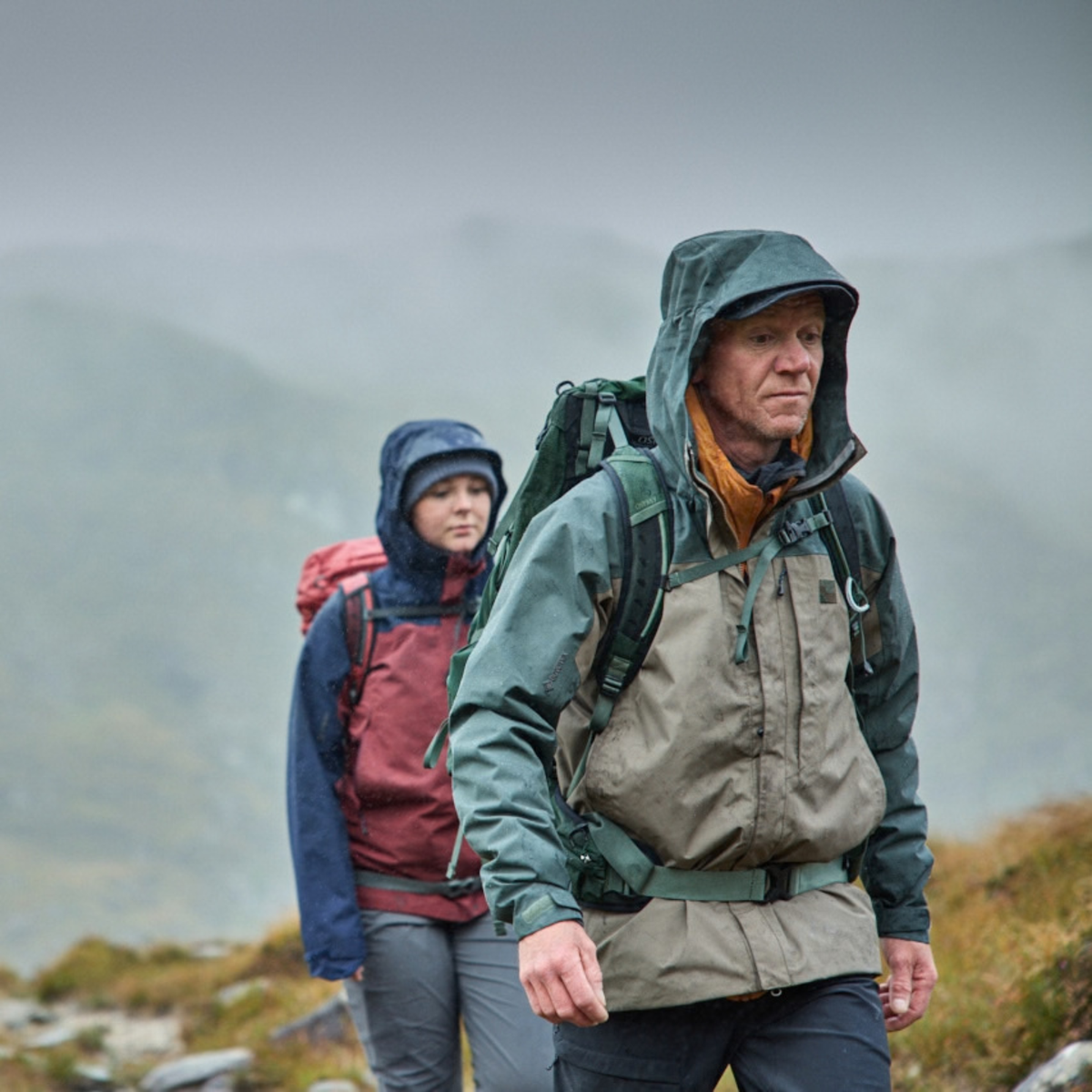 Two people hiking outdoors in the rain, Man wearing Sprayway Men's Reaction 2L Gore-Tex Jacket SP-01723 Brownstone Bonsai SP-008036 and carrying a backpack on a misty mountain trail.