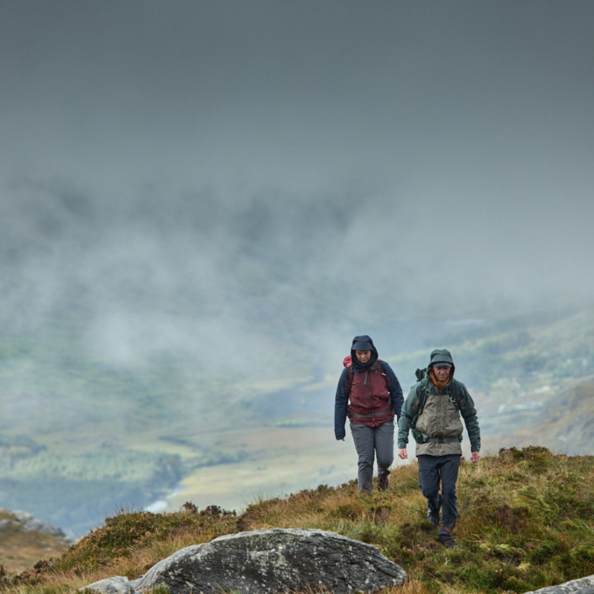 Two hikers walking along a grassy mountain ridge under a dramatic, cloudy sky with misty valleys in the background. Man wearing Sprayway Men's Reaction 2L Gore-Tex Jacket SP-01723 Brownstone Bonsai SP-008036