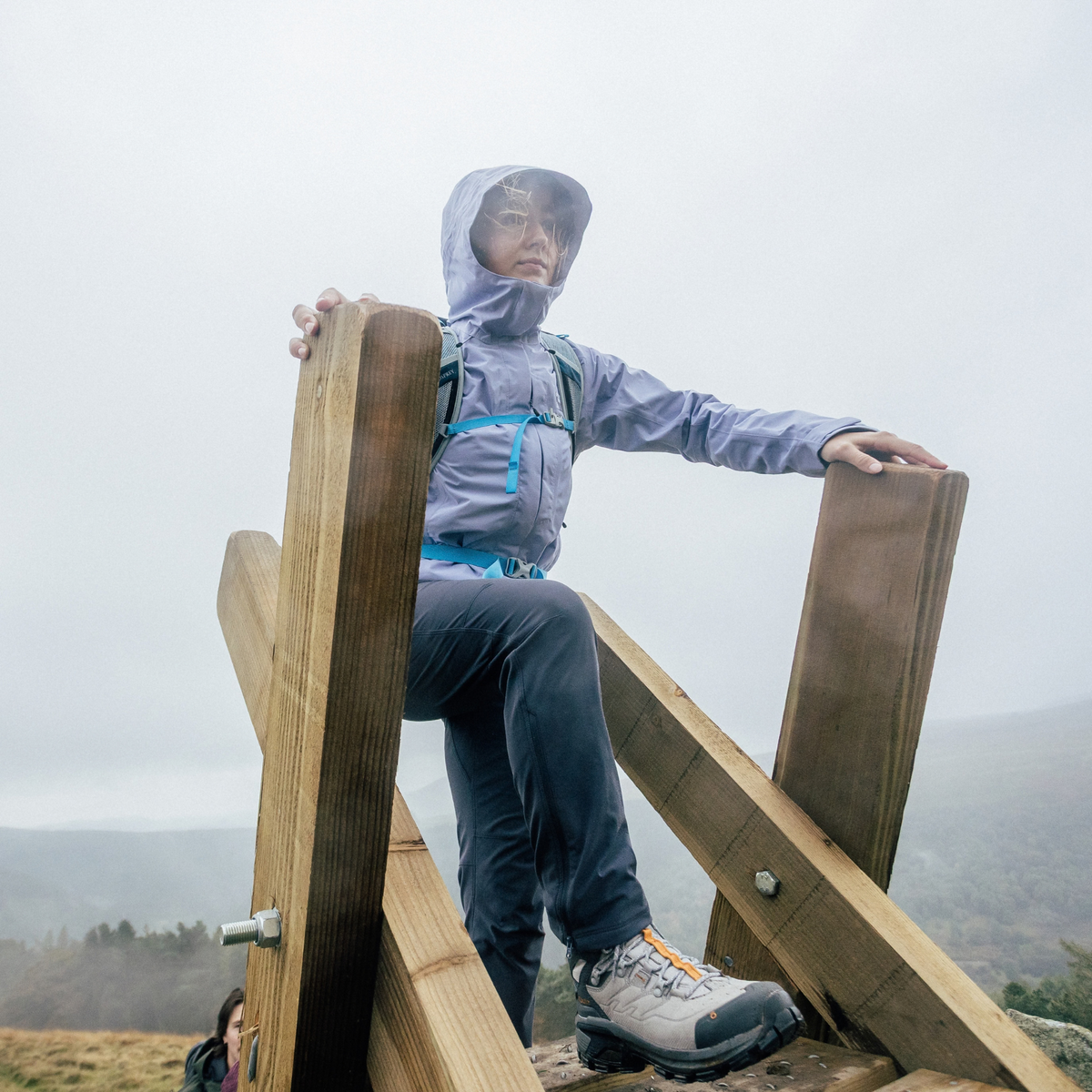 A person wearing a lilac hooded rain jacket and Sprayway Women's Escape Softshell Warm Walking Hiking Trousers SP-01006 Black SP-007510 climbing over a wooden turnstyle outdoors in the rain, with a misty mountain landscape in the background.