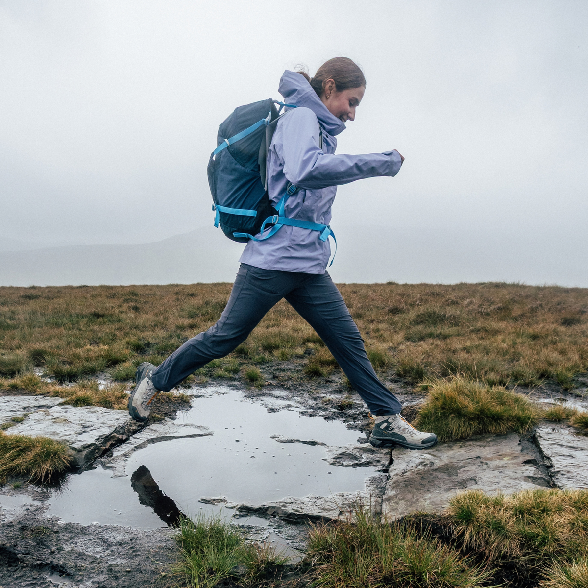 A woman hiking on a muddy, overcast mountain path wearing a light purple waterproof jacket, Sprayway Women's Escape Softshell Warm Walking Hiking Trousers SP-01006 Black SP-007510, and a blue hiking backpack.