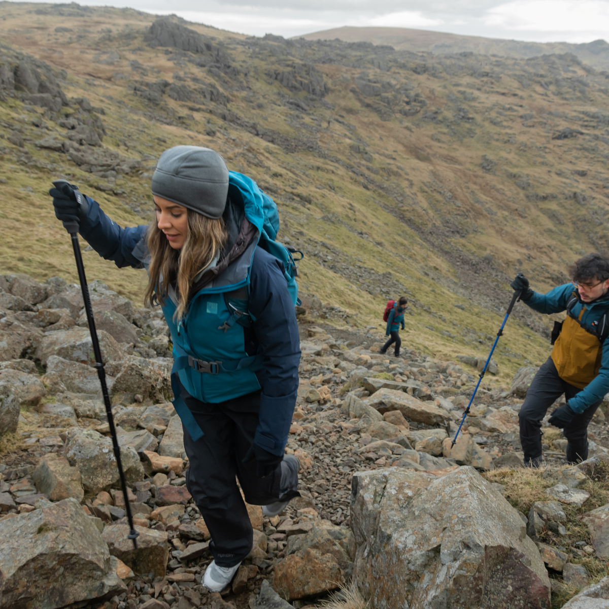 Two hikers climbing a rocky mountain with another person in the background. Woman using Trekmates Hiker Shock Single Pole Black TM-008705