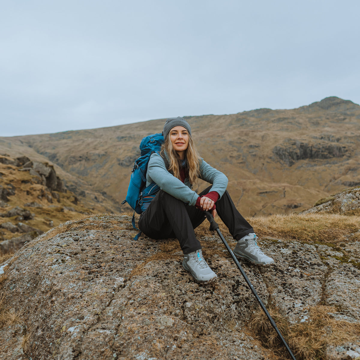 Person with a backpack sitting on a rocky outcrop in a mountainous landscape holding Trekmates Hiker Shock Single Pole Black TM-008705