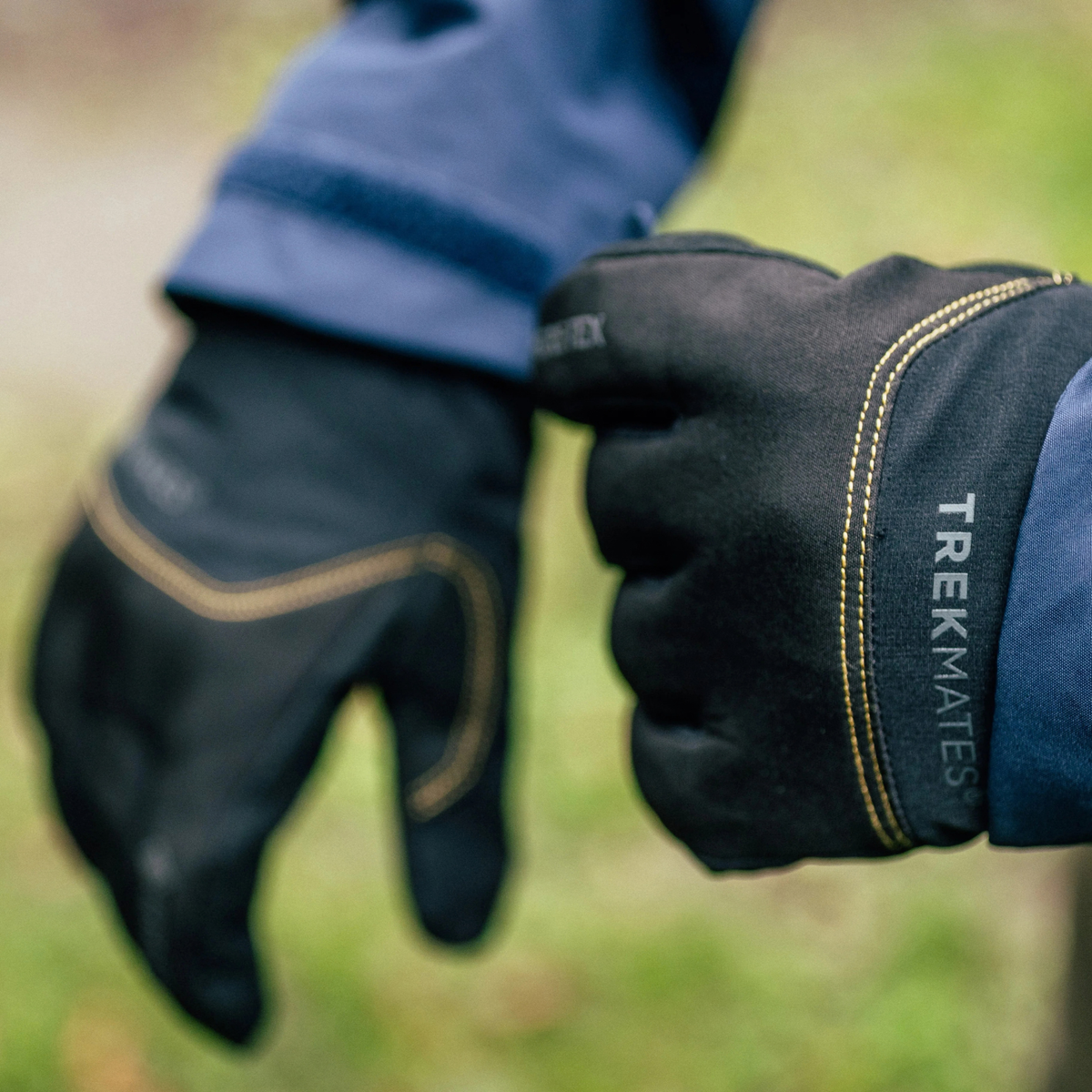 Trekmates Unisex Friktion Gore-Tex Waterproof Gloves Black TM-008647 shown being worn with a blue jacket sleeve, photographed outdoors with a blurred green background.