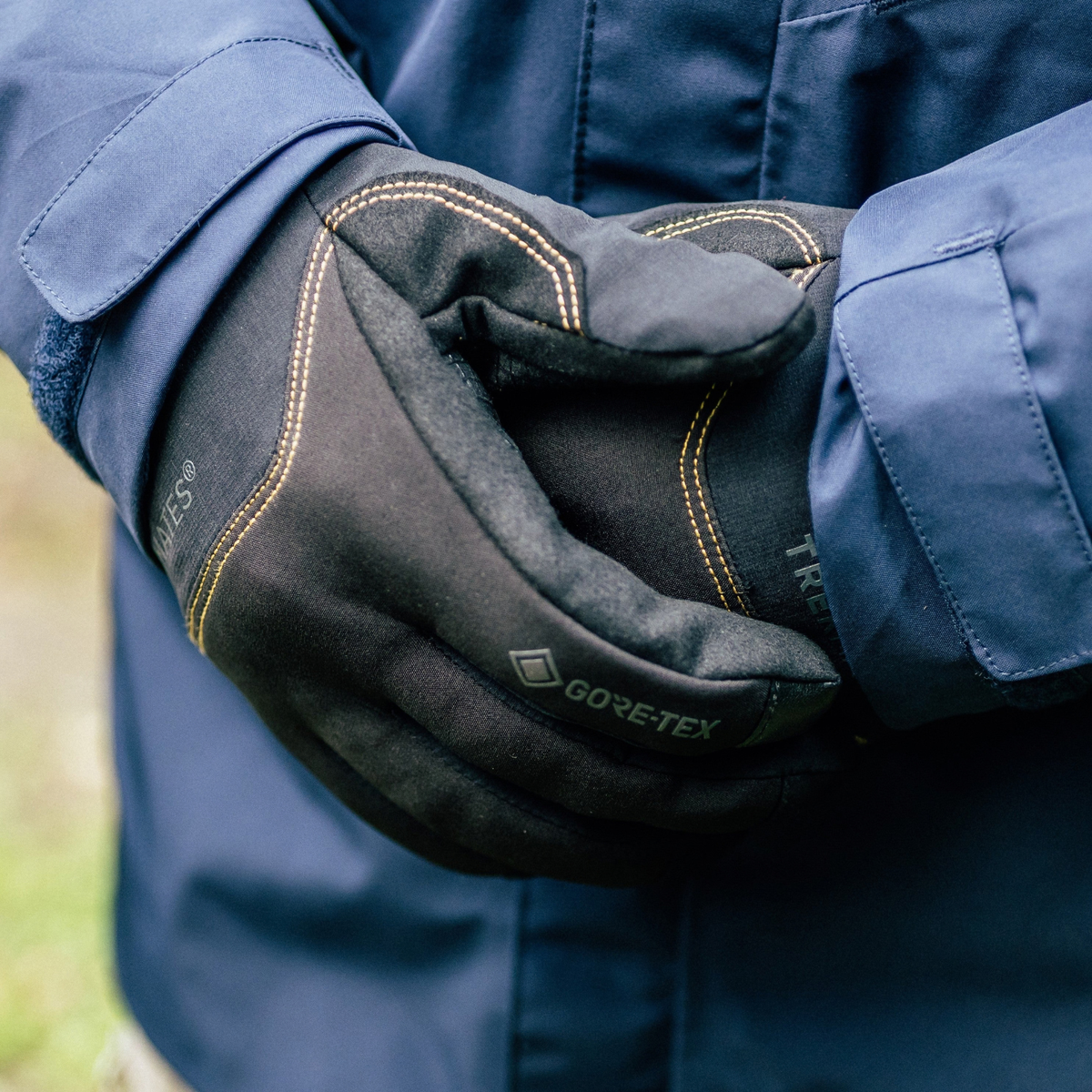Close-up of a person wearing Trekmates Unisex Friktion Gore-Tex Waterproof Gloves Black TM-008647 over a navy blue jacket, with the hands folded over to show the glove construction and stitching.