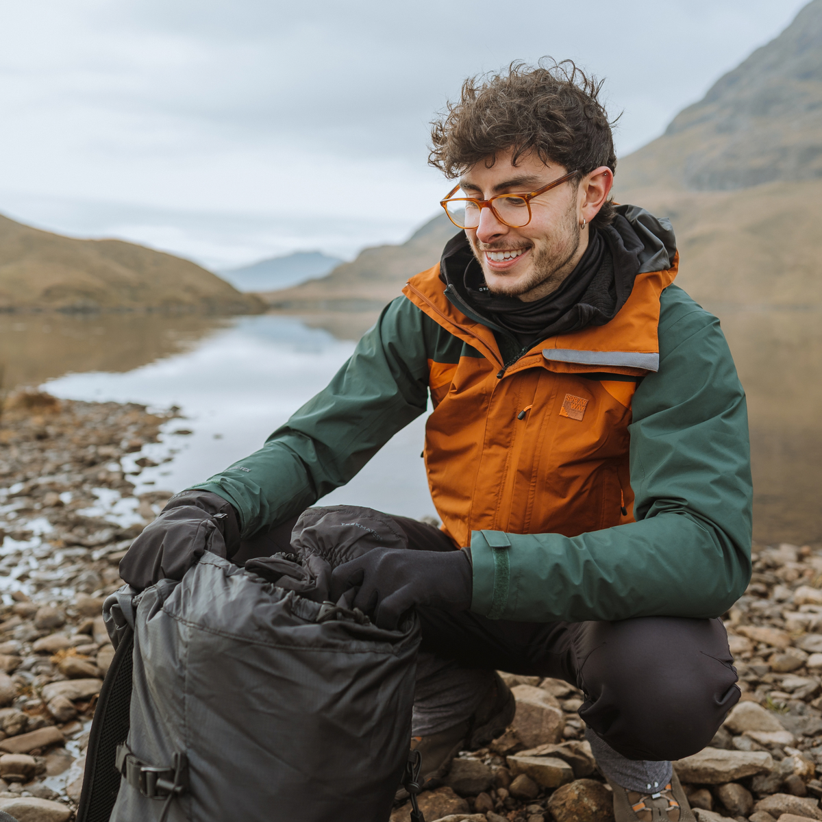 Man wearing Trekmates Unisex Silk Touch Liner Gloves Black GLV-TH-U10547 and waterproof jacket, kneeling down on stony ground beside a lake up the mountains.