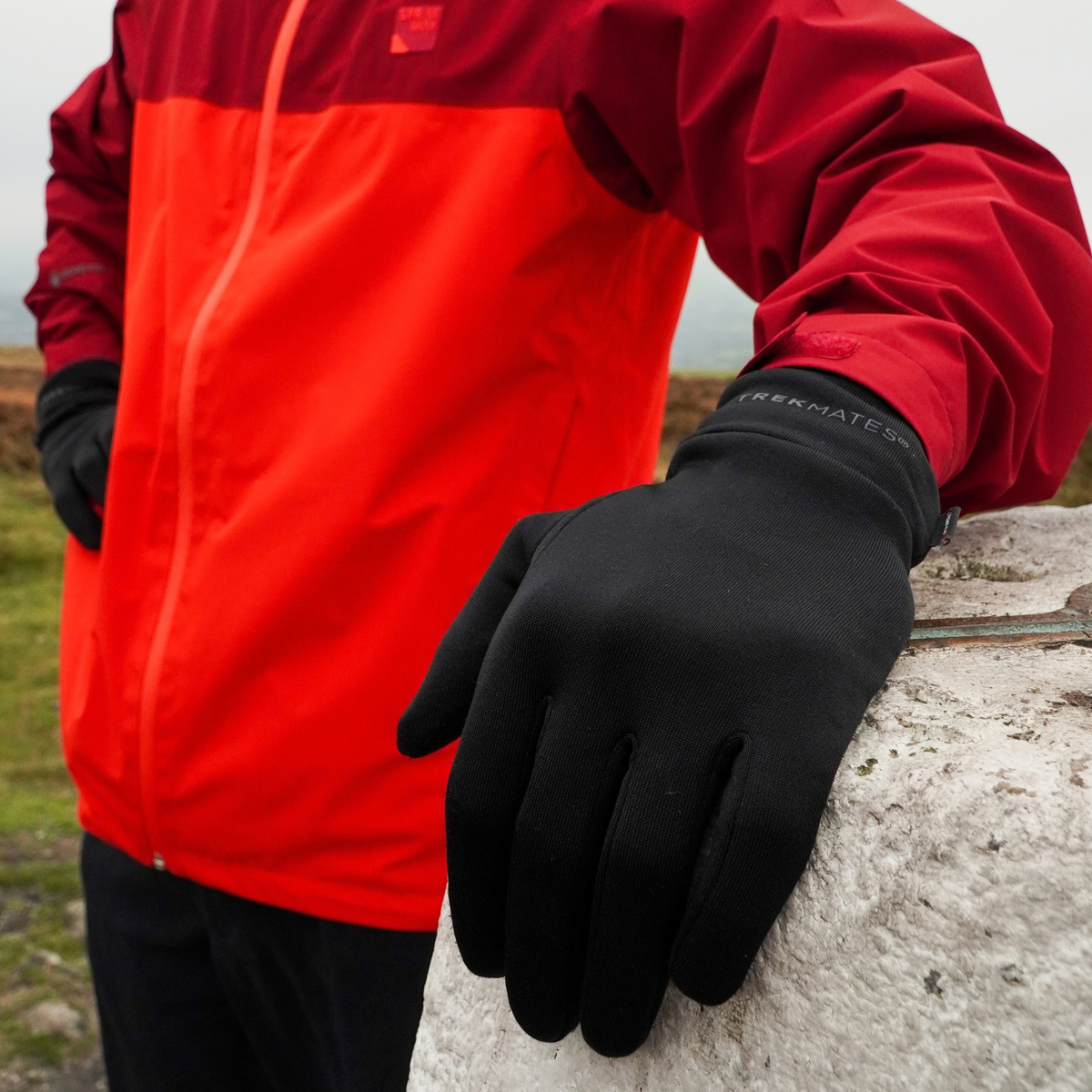 Man wearing Trekmates Unisex Silk Touch Liner Gloves Black GLV-TH-U10547 with his hand placed on a summit marker on top of a mountain