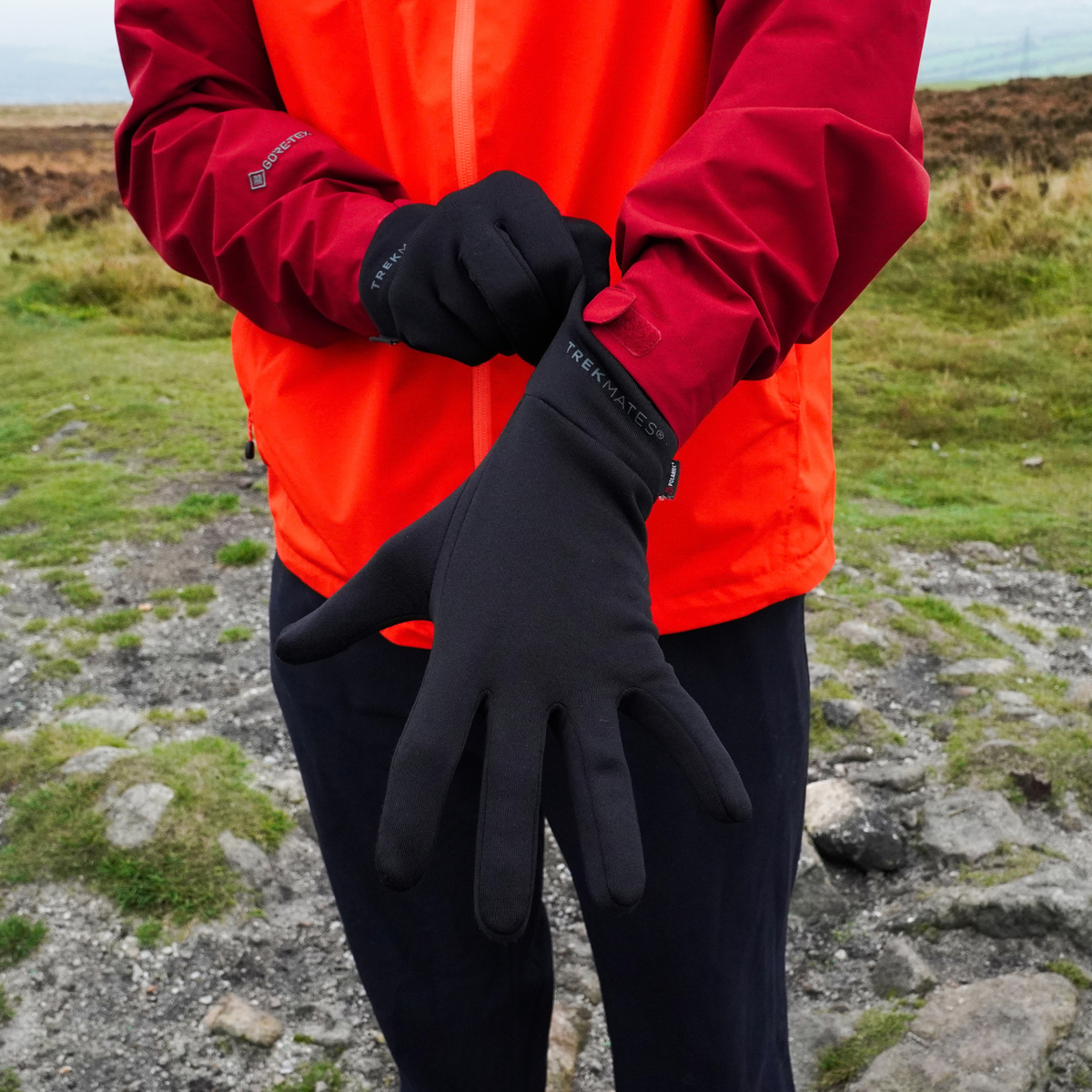 Man pulling on Trekmates Unisex Silk Touch Liner Gloves Black GLV-TH-U10547 wearing a red waterproof jacket with a grassy background