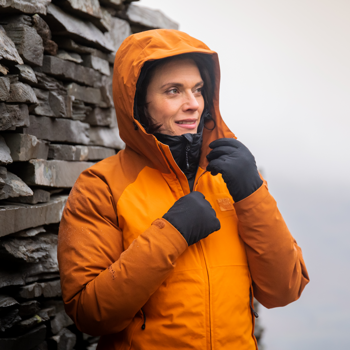 Person wearing an orange outdoor waterproof jacket and Trekmates Women's Bala DRY Waterproof Gloves Black TM-008785 standing against a stone wall.