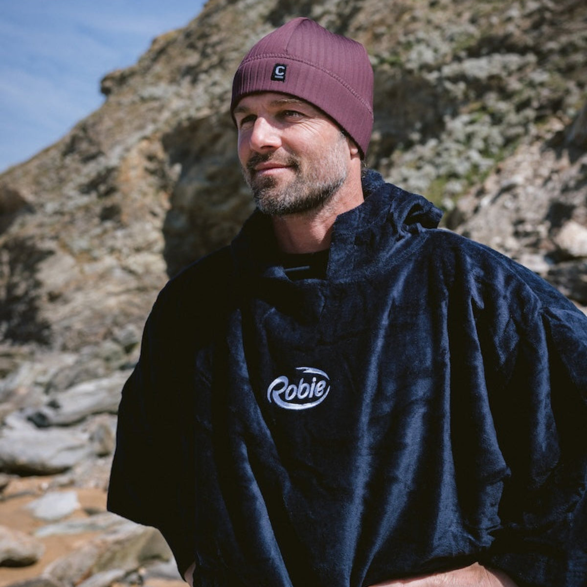 Man wearing a C-Skins Unisex Storm Chaser Thermal Neoprene Swim Watersports Beanie Rust C-HOBE and black Robie changing robe, standing outdoors against a blue sky.