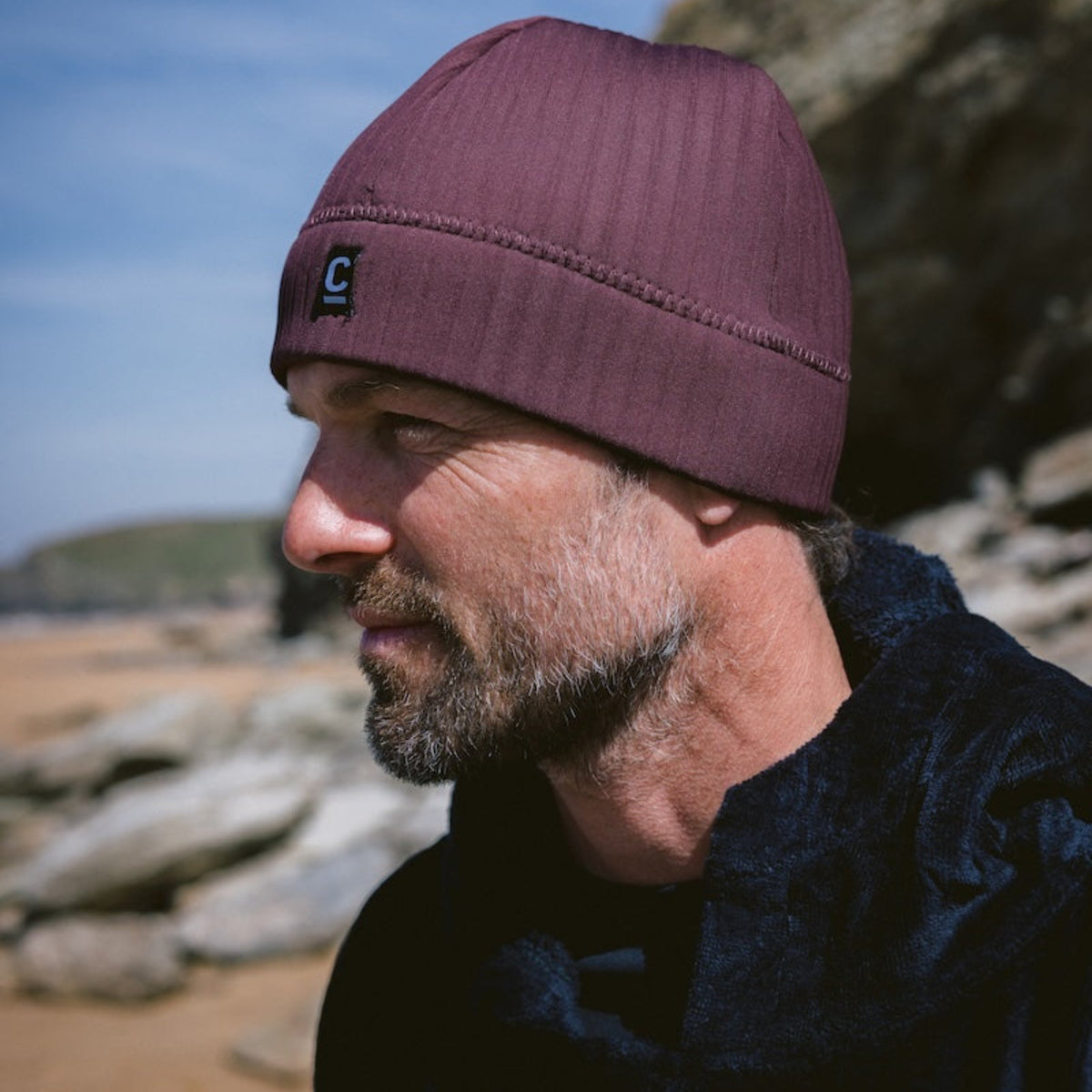 Man wearing a C-Skins Unisex Storm Chaser Thermal Neoprene Swim Watersports Beanie Rust C-HOBE and black Robie changing robe, standing outdoors against a blue sky.