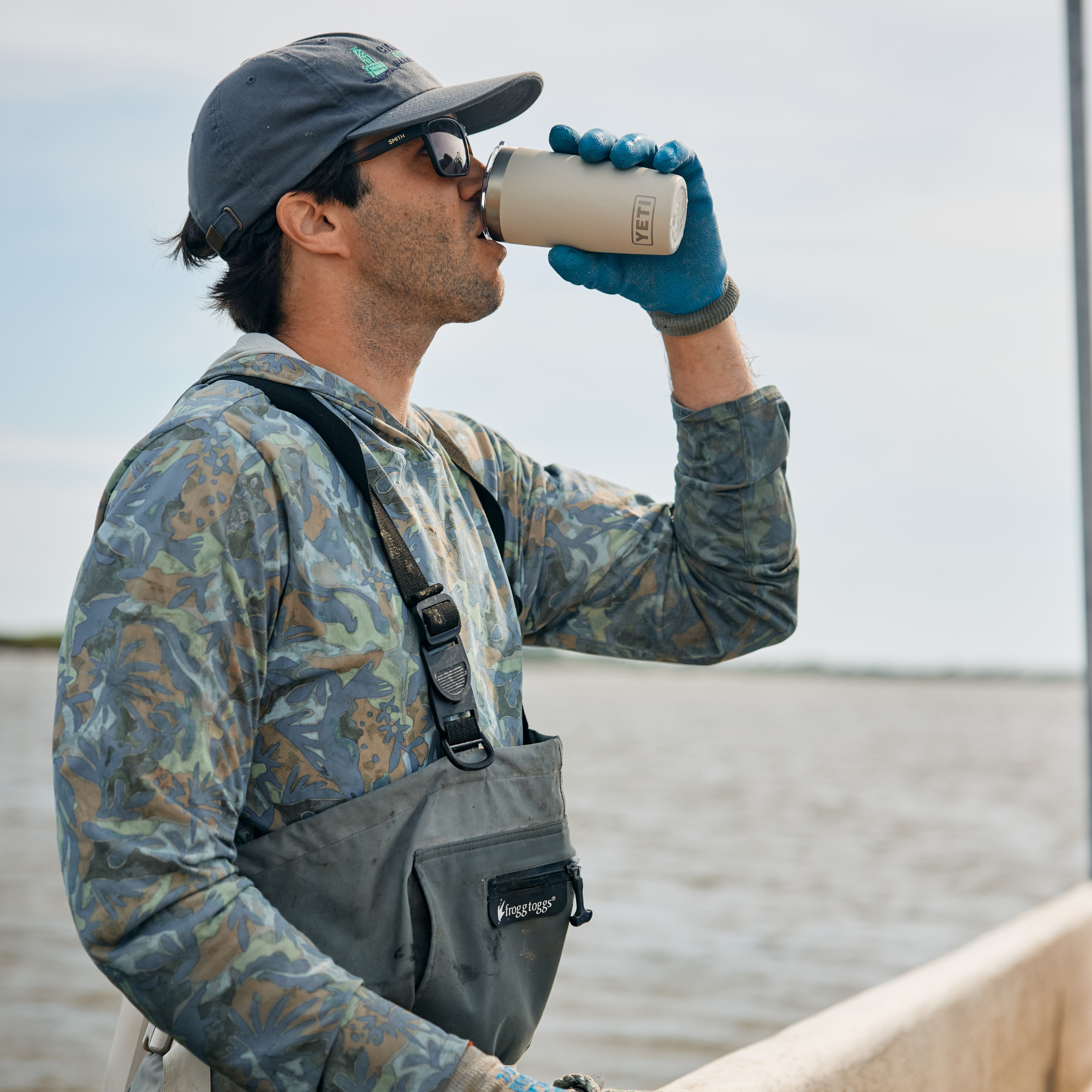 A man wearing a camouflage long-sleeve shirt, grey waders, and a dark cap drinks from a Yeti Rambler 10oz Tumbler Cape Taupe 70000003165 Yeti-RamblerTumbler-10oz while standing outdoors near water.