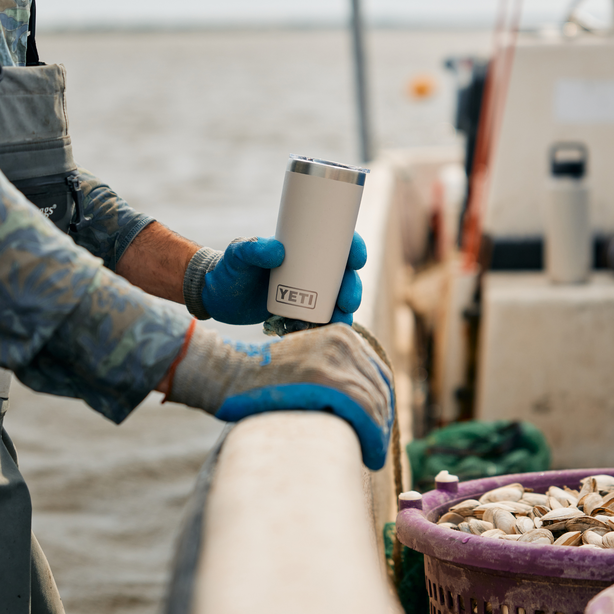 A person on a boat wearing blue gloves holds a Yeti Rambler 10oz Tumbler Cape Taupe 70000003165 Yeti-RamblerTumbler-10oz. The background shows the ocean water and a fishing boat deck, with a purple basket of clams in the foreground.