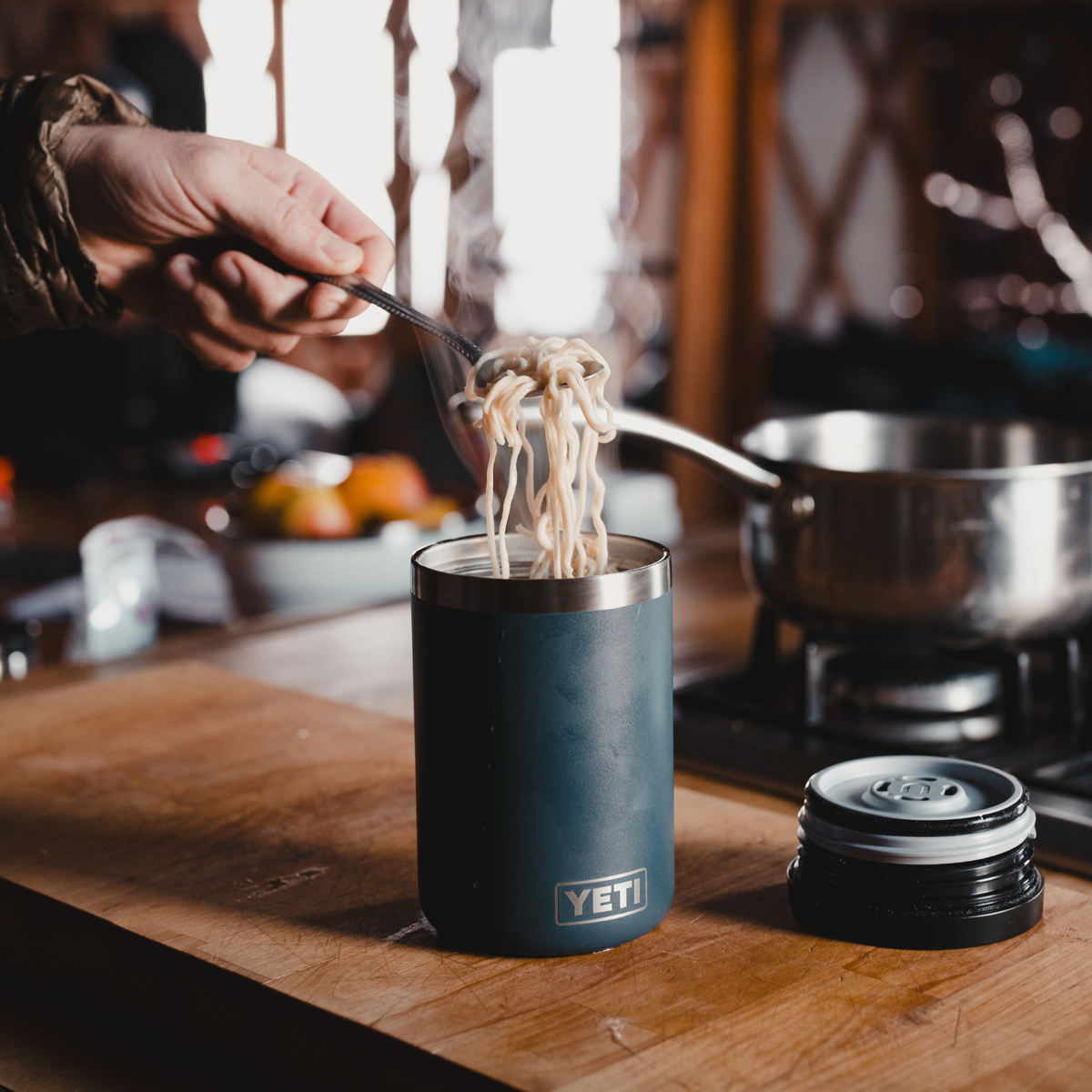 Yeti Rambler Insulated Food Jar is shown on a wooden countertop in a kitchen setting, with a hand using a fork to lift noodles from the open top of the tumbler. The lid sits beside it.