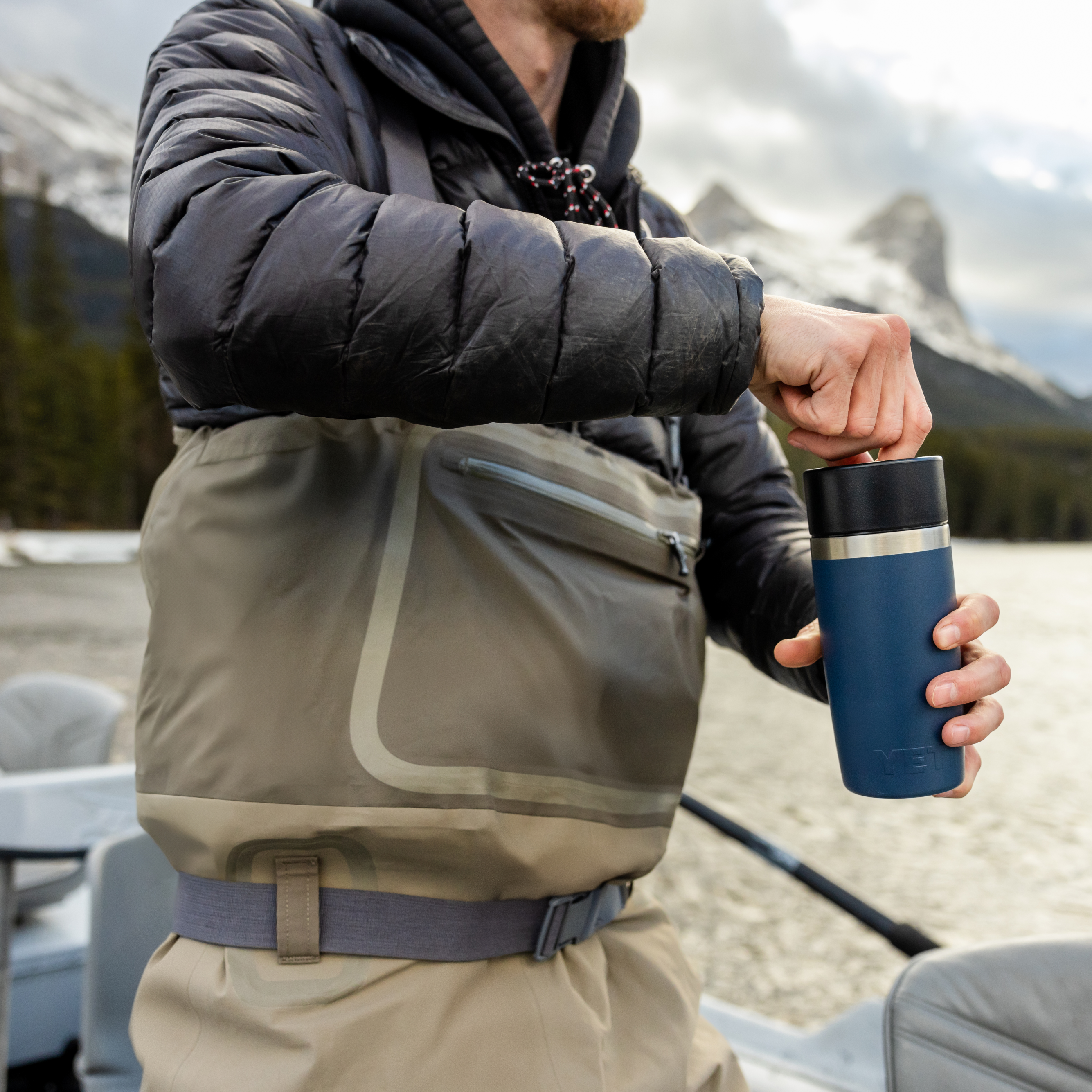 Man holding a navy Yeti travel bottle with commuter lid on a fishing boat wearing fishing waders