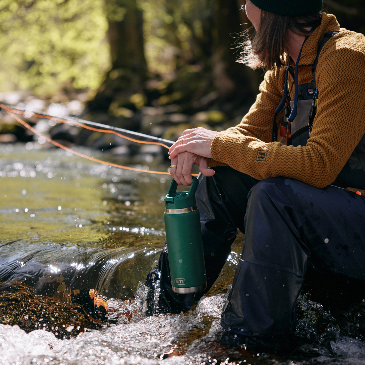 A person in a forest stream holds a Yeti Rambler 26oz Insulated Bottle Straw Cap Black Forest Green 70000004236 Yeti-RamblerStraw-26oz while wearing a mustard yellow knit sweater, dark waders, and a black beanie.