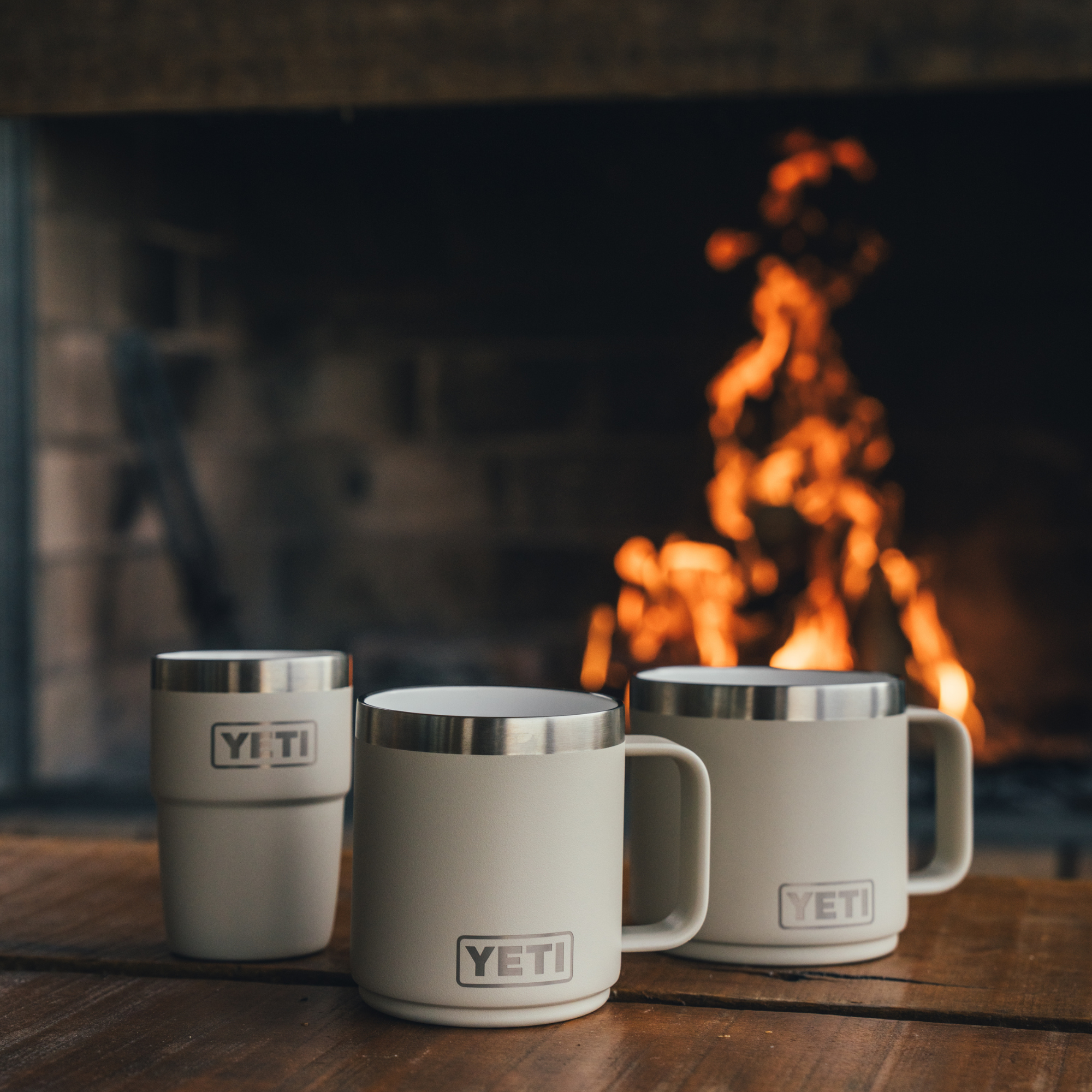 Three cream Cape Taupe Yeti Travel Mugs arranged on a wooden table with a fireplace blurred background.