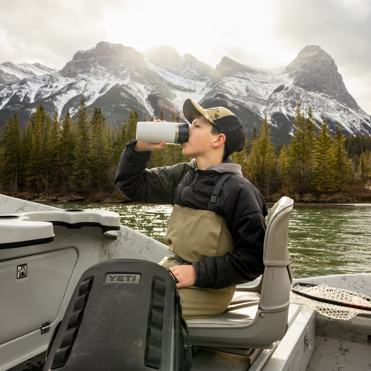 Person sitting in a boat on a lake with mountains in the background drinking out of a yeti rambler insulated travel mug