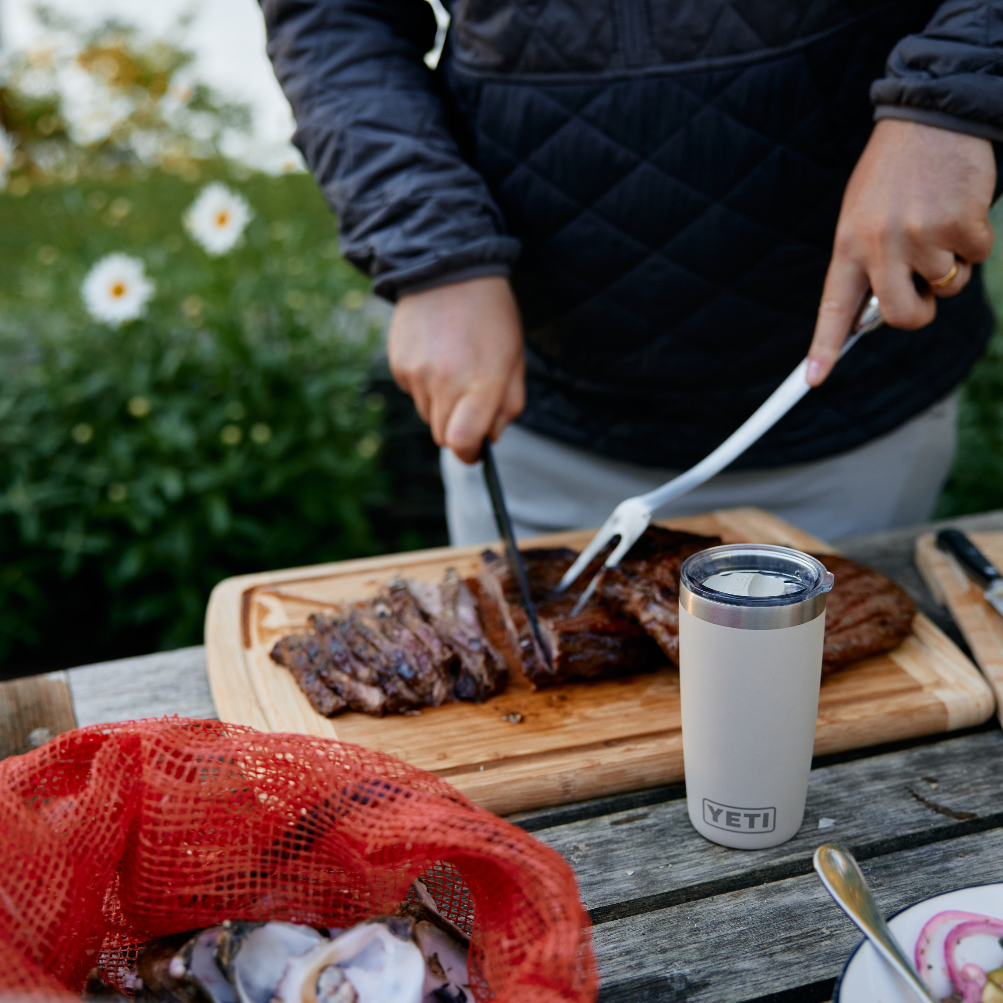 A person outdoors at a wooden table slices a grilled steak on a cutting board, with a Yeti Rambler Tumbler Insulated Travel Mug Magslider Lid 20oz 591ml Cape Taupe 70000003166 Yeti-RamblerTumbler-20oz, a red mesh bag, and a plate with sliced onions nearby.