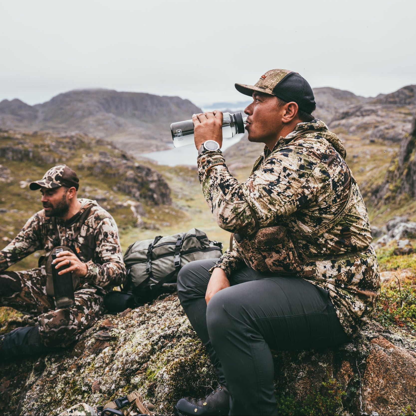 Two individuals in camouflage attire sitting on a rocky mountain landscape, one drinking from a Yeti Yonder 34oz 1L Water Bottle with Tether Cap Charcoal 70000002374 Yeti-YonderBottle-34oz
