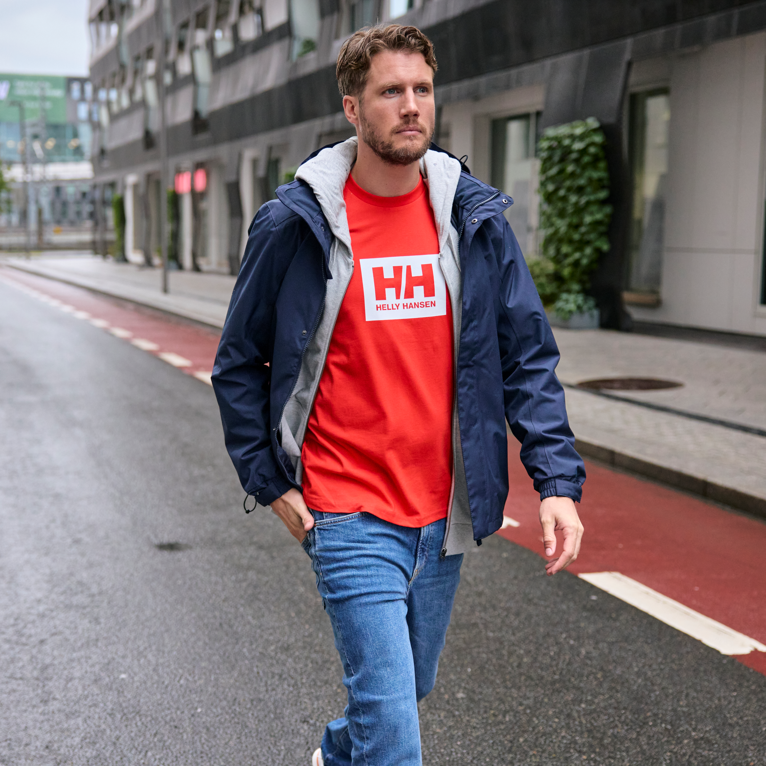 Man walking in a city wearing an orange Helly Hansen t-shirt and an open 62643 Helly Hansen Men's Dubliner Waterproof Rain Jacket Navy 597 with water droplets on the jacket